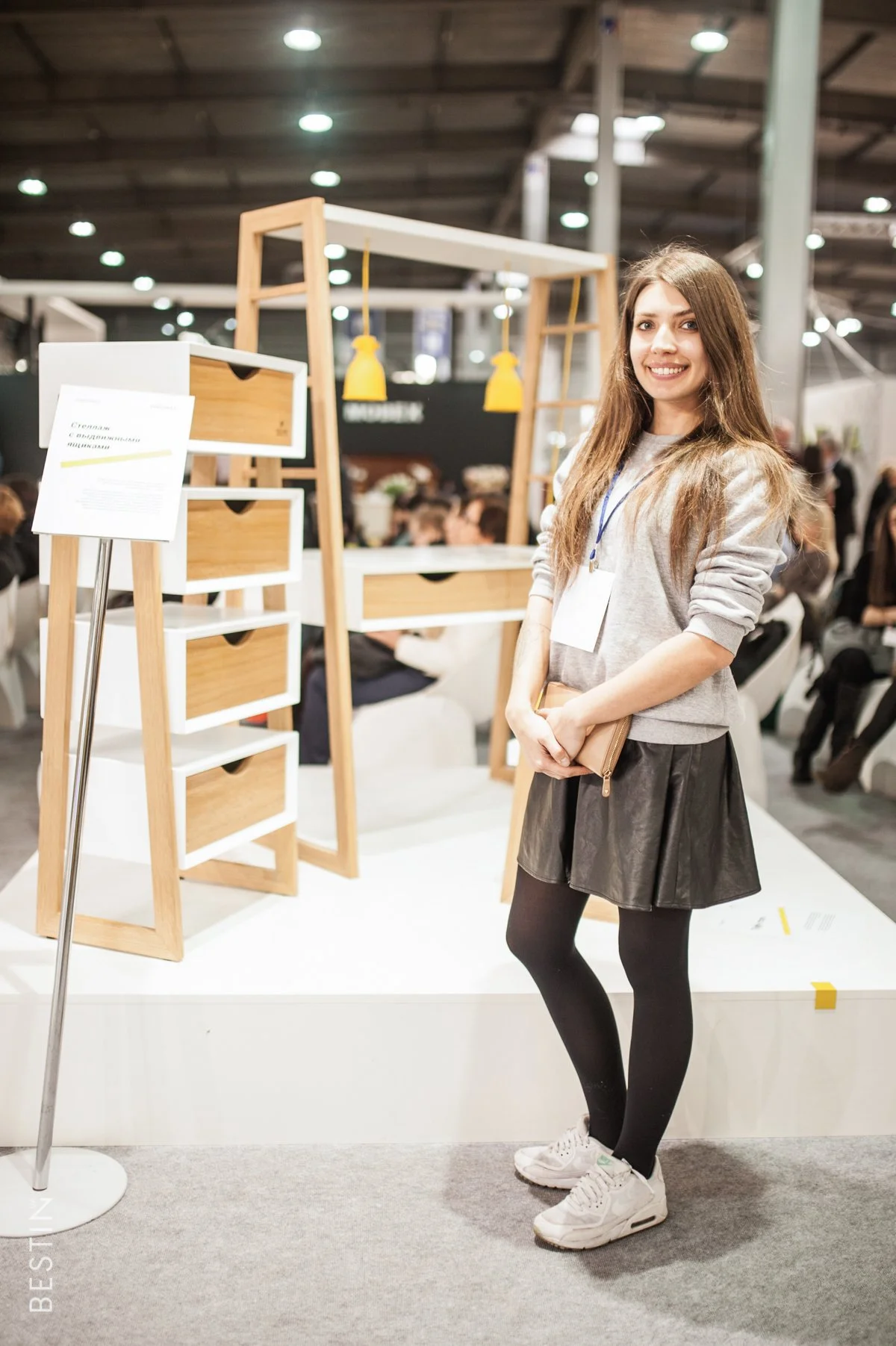 A young woman with long brown hair standing at a trade show booth exhibiting wooden furniture, smiling and holding a beige clutch bag.
