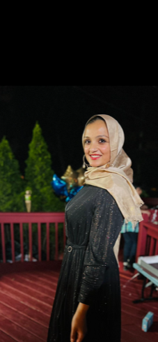 A woman wearing a beige headscarf and black dress, smiling outdoors at night with string lights and greenery in the background.