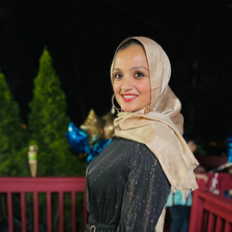 A woman wearing a beige headscarf and black dress, smiling outdoors with greenery and colorful balloons in the background.