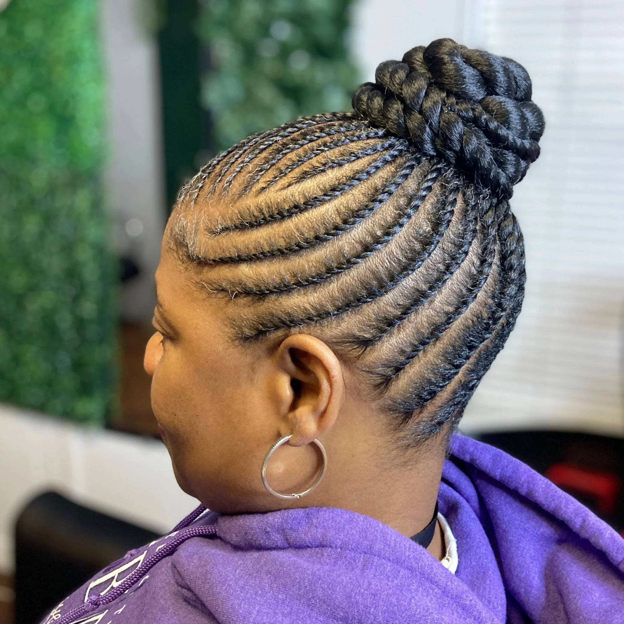 A woman with braided hair styled in a high bun, wearing a purple hoodie and hoop earrings.