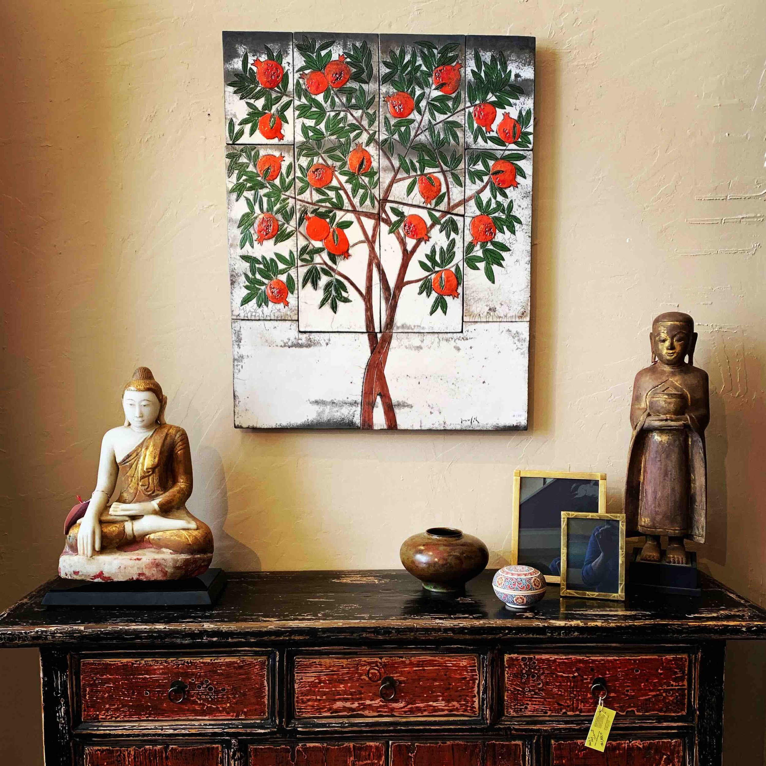 Decorative indoor wall art featuring a pomegranate tree with orange-red fruit and green leaves, painted on a triptych panel. The artwork is displayed above a wooden table with various objects, including statues and picture frames.