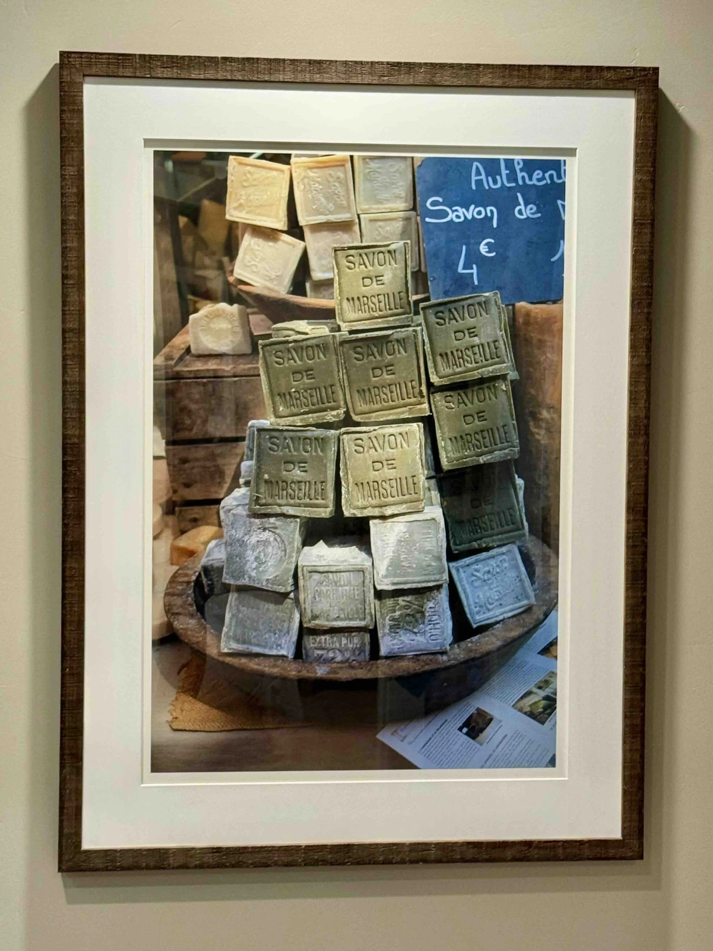 Framed photograph of an assortment of soaps labeled 'Savon de Marseille' stacked on a wooden surface with a blue sign showing the price of 4 euros.