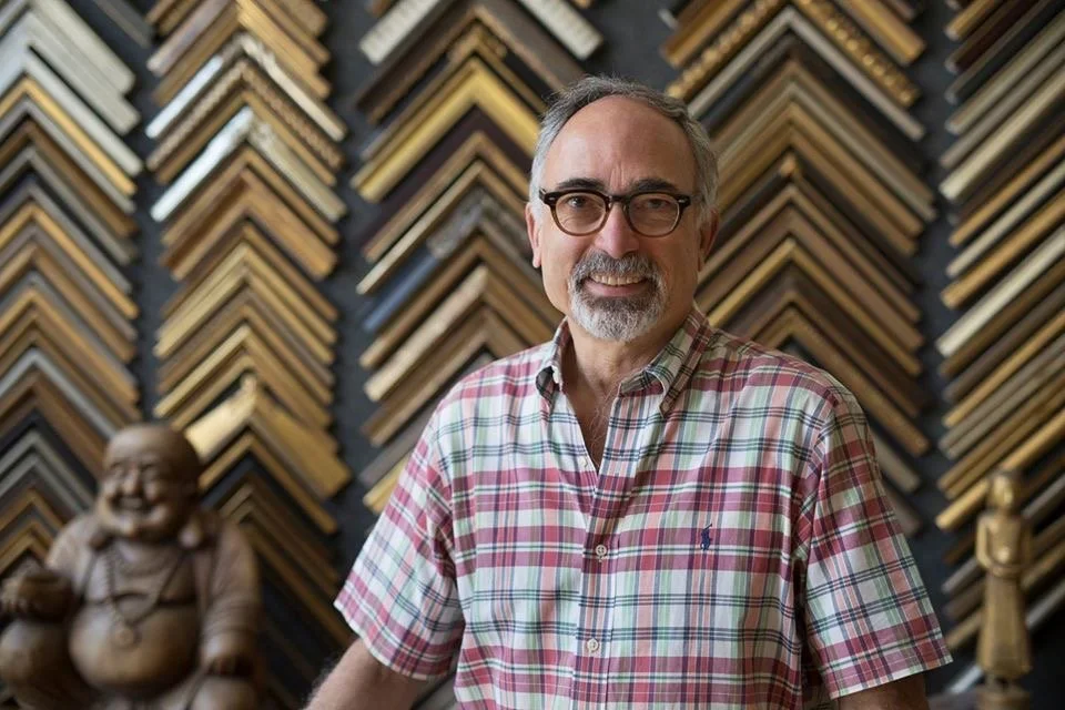 Proprietor and designer of picture framing shop, standing in front of a wall decorated with frame samples.