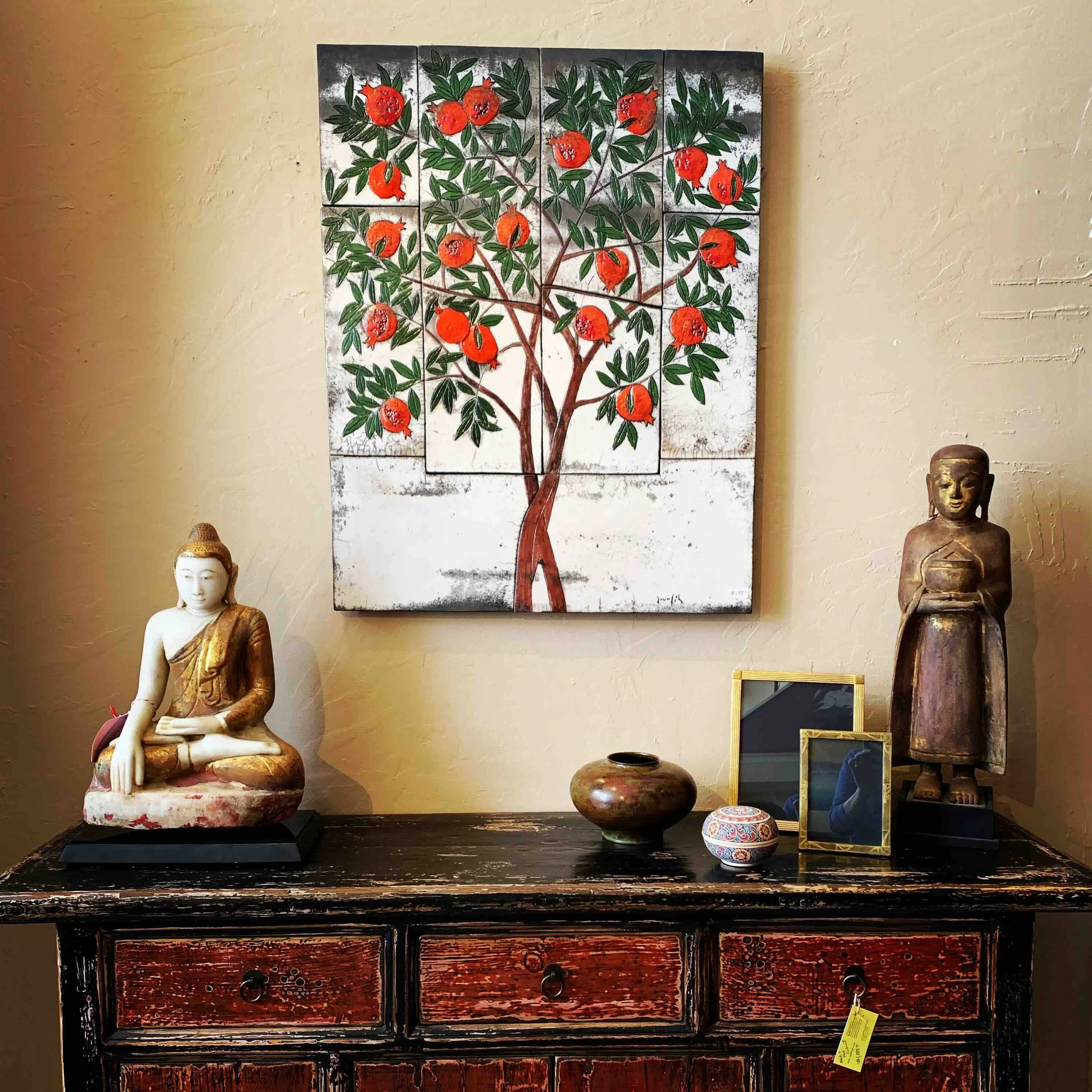 A multi-panel ceramic artwork of a pomegranate tree with green leaves and red fruit, hung on a wall behind an antique Chinese console table with various decorative objects, including a marble Buddha, a bronze bowl and desktop picture frames.