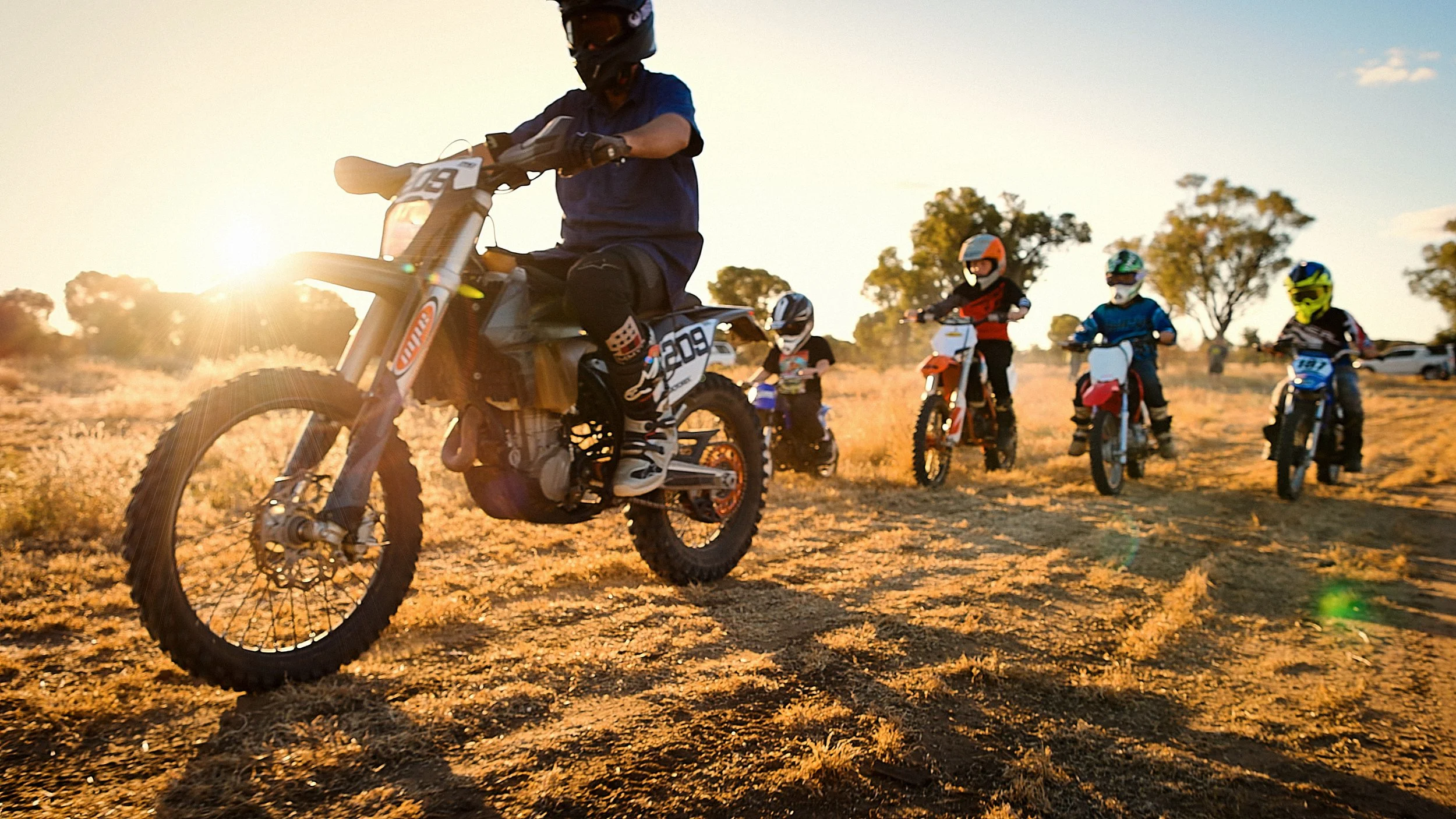 A group of children and a man riding dirt bikes on a dirt trail during sunset, wearing helmets and protective gear.