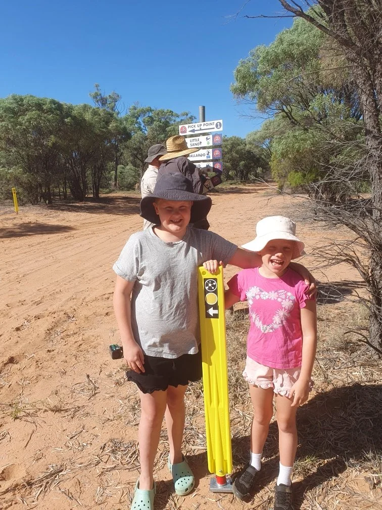 Two children standing on a dry dirt trail, smiling, with trees and a blue sky in the background. They are wearing hats and casual clothing, and there is a bright yellow surveying tool between them. In the background, a signpost indicates directions to various locations.