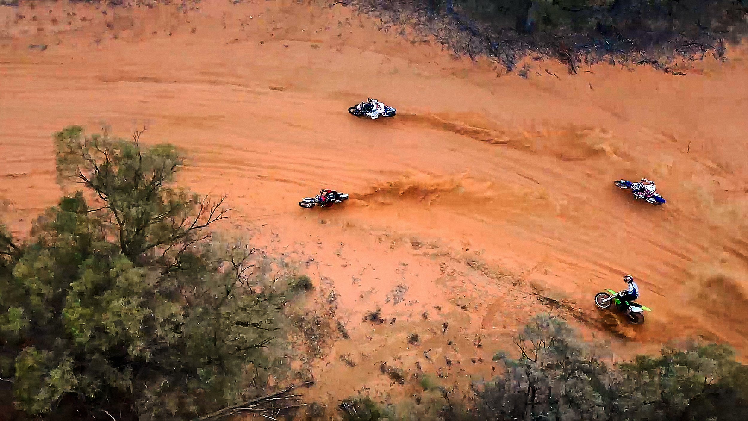 Five motorcyclists riding on a dusty, reddish-brown dirt trail through a semi-arid landscape with sparse trees.