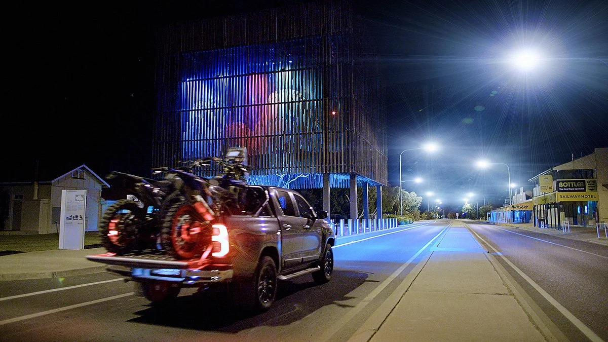 A pickup truck is parked on the side of a city street at night, carrying two motorcycles in its bed. The street is empty, illuminated by streetlights, with a modern building with colored lights in the background.