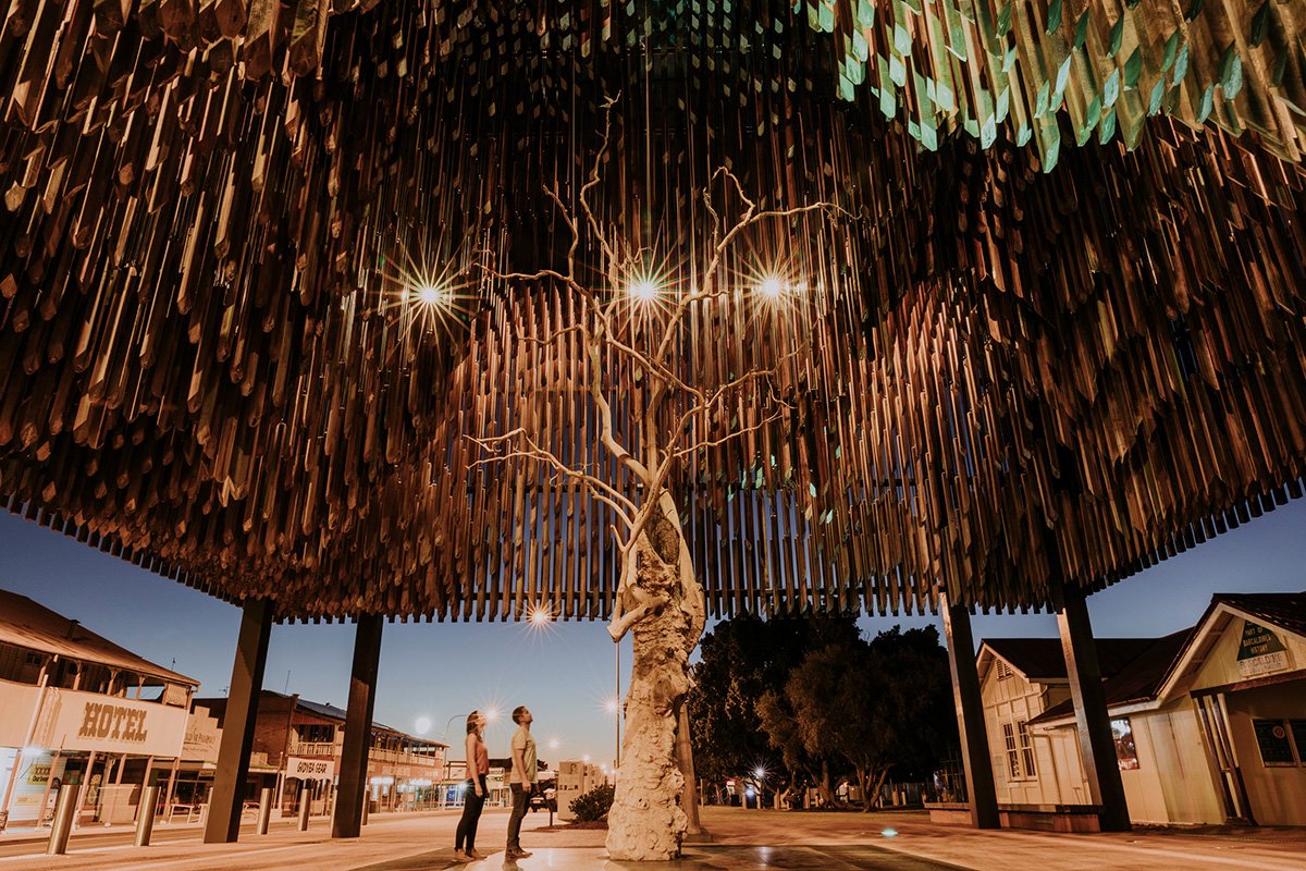 Three people standing under a large artistic canopy with a tree sculpture in the center and overhead lights at night.