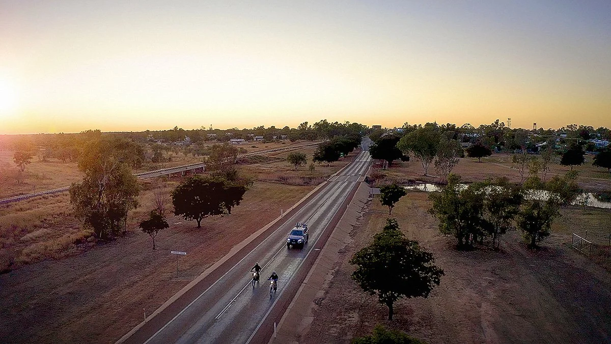 An aerial view of a rural road at sunset with trees, a bike lane, a car, and two cyclists.