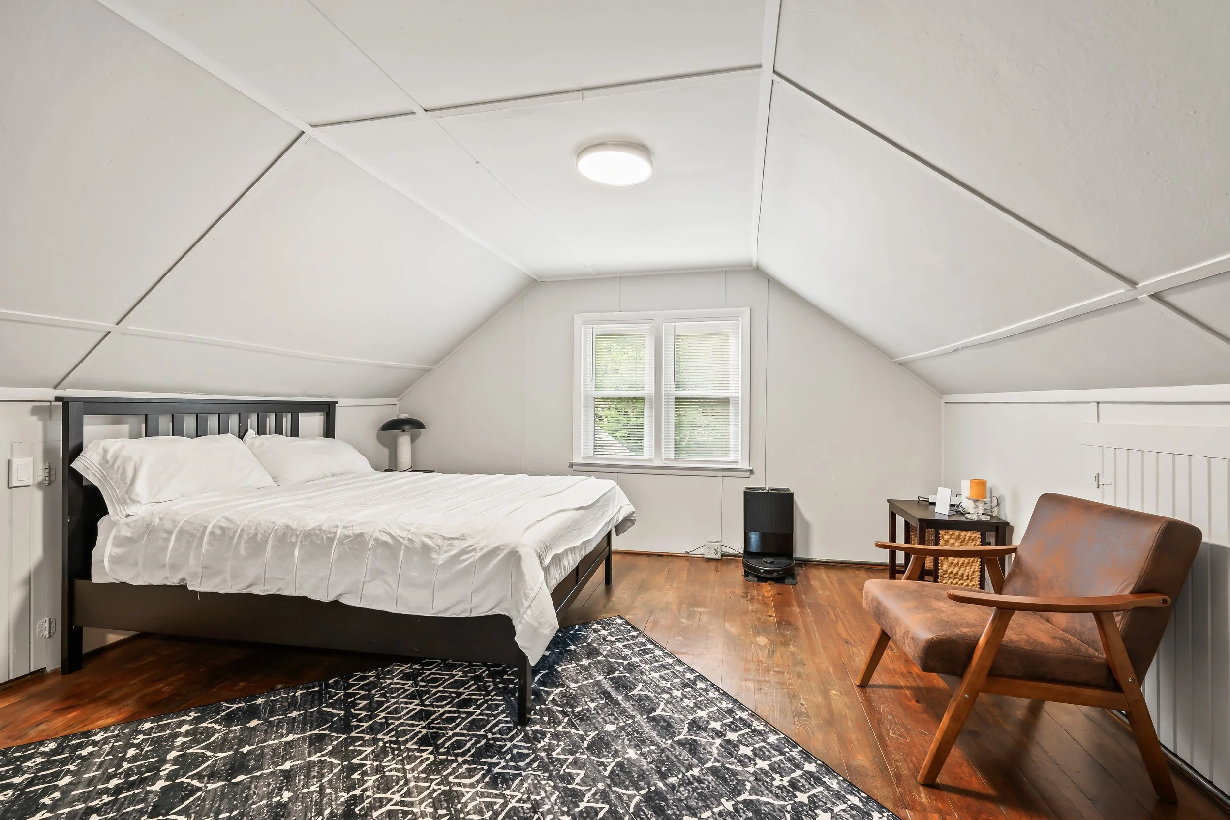 Bedroom with white ceiling, window, black bed frame, white bedding, black nightstand with lamp, brown armchair, and black patterned rug on wooden floor.