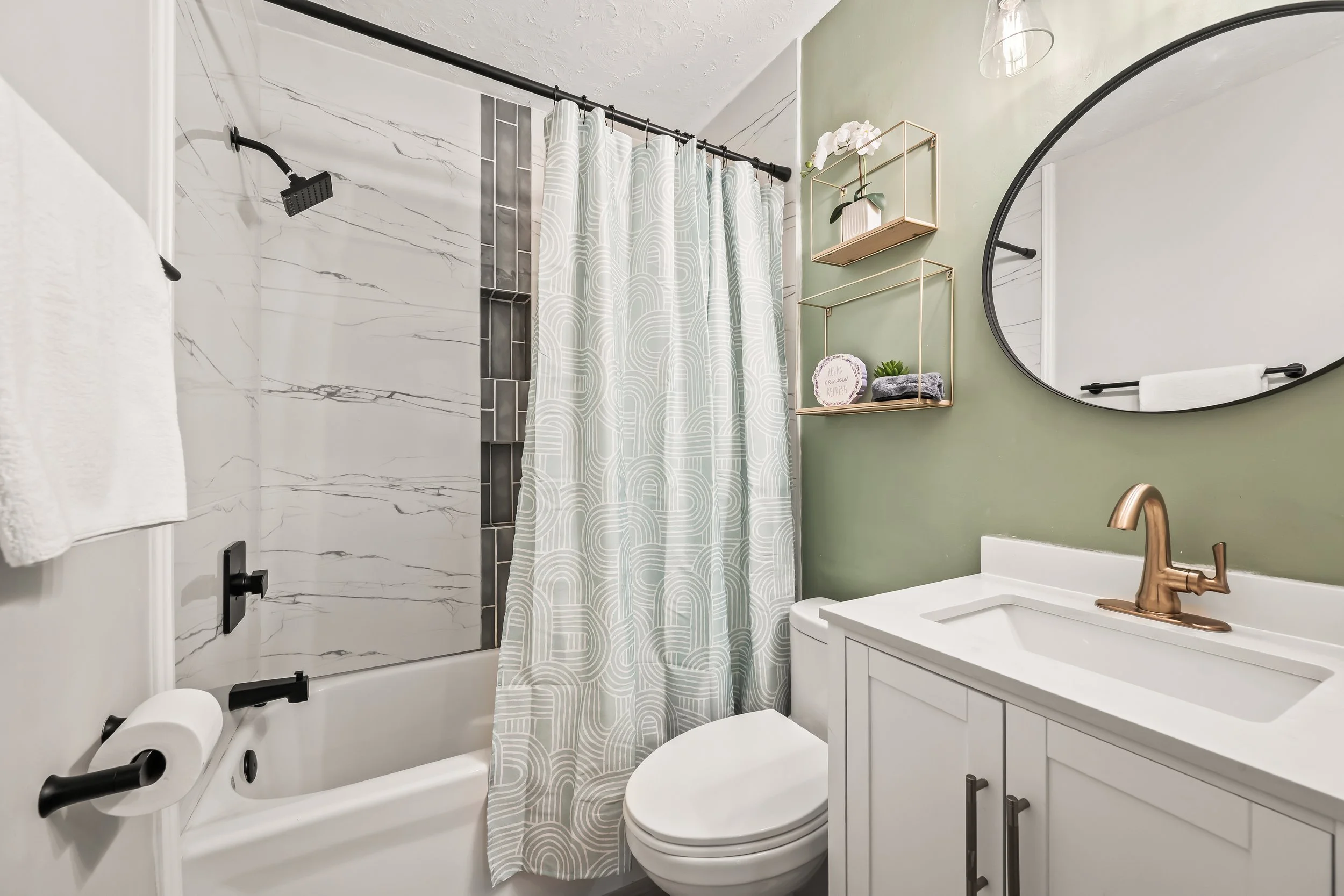 A bathroom with a shower area with marble tile walls, a curtain with geometric patterns, a round mirror on a green wall, a floating shelf with a flower and decor, a white vanity with a sink and copper faucet, and a toilet.