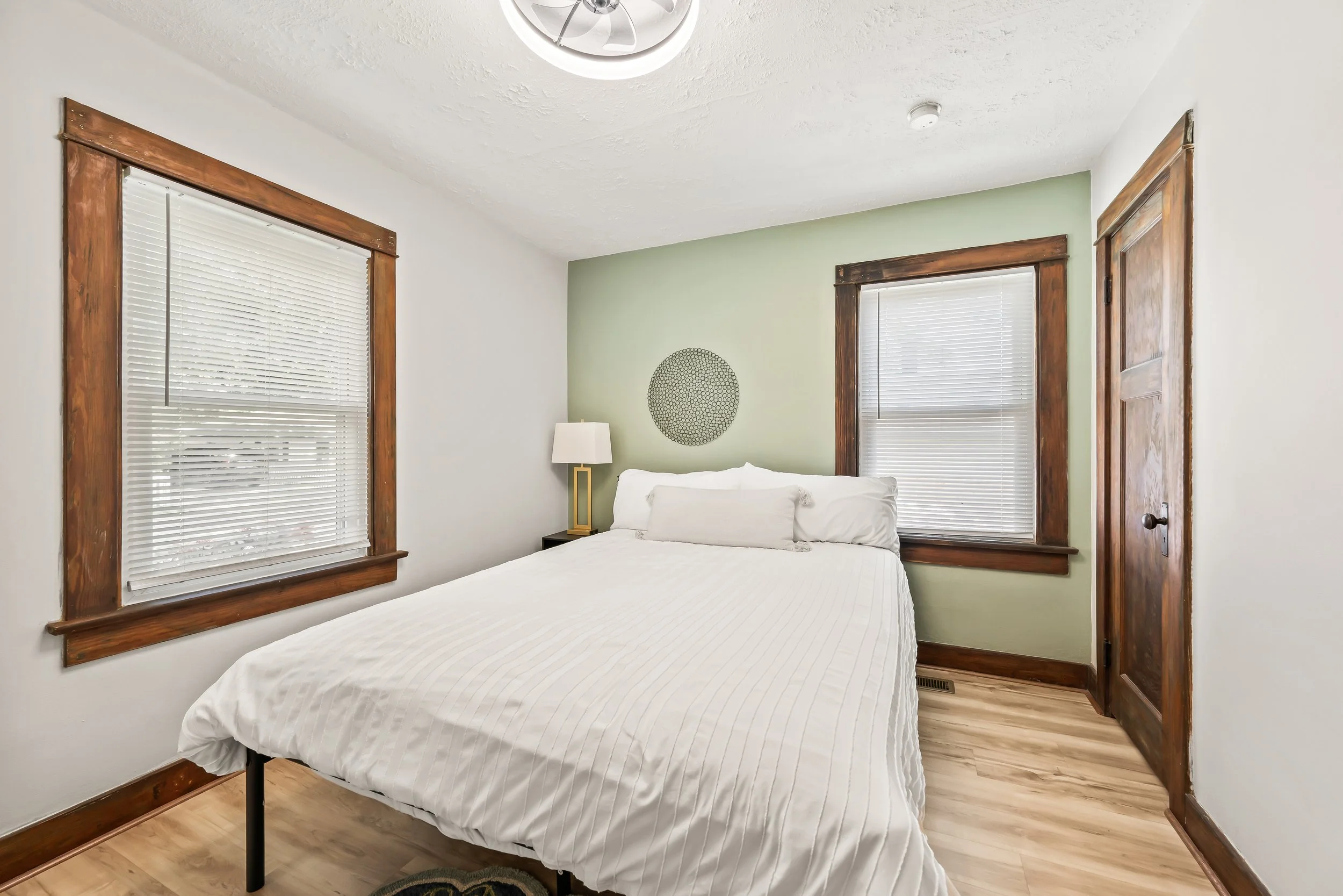 A bedroom with a bed covered in a white bedspread, two large windows with white blinds and wooden frames, a small nightstand with a lamp, and a light fixture on the ceiling.