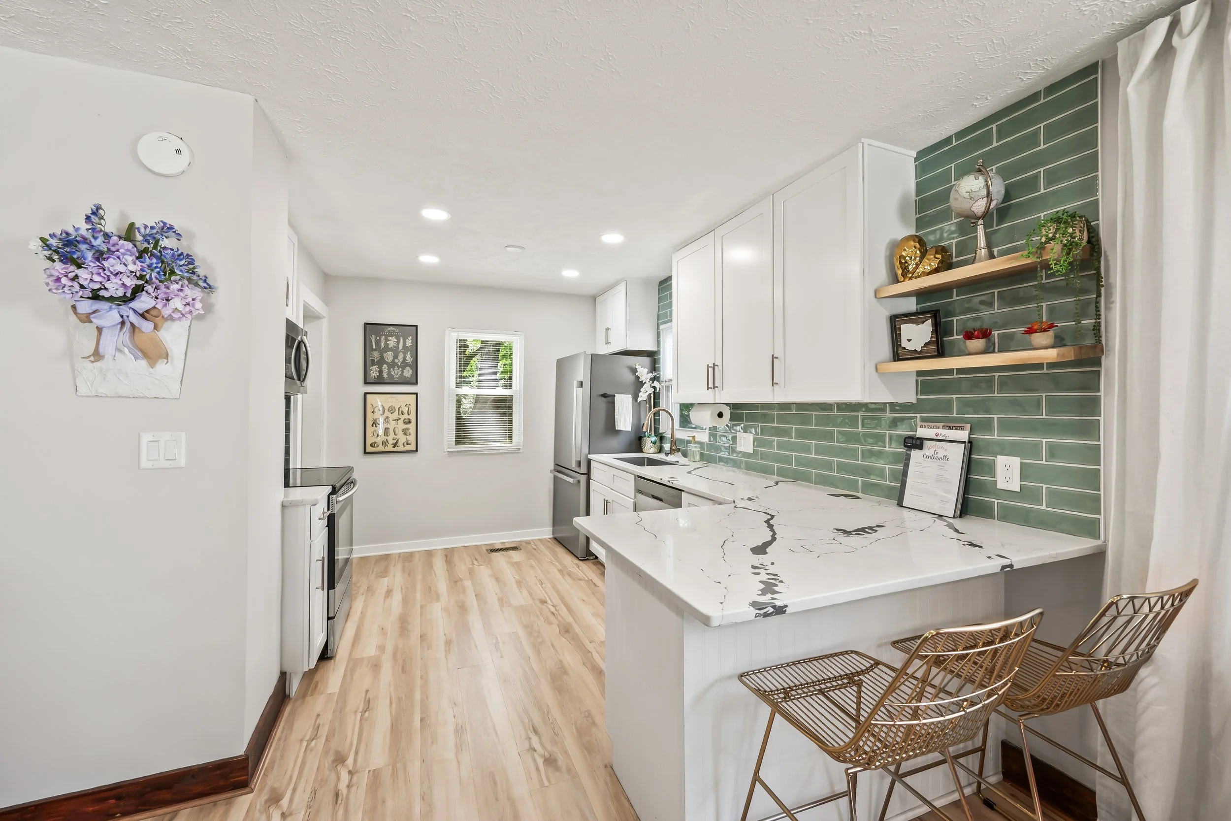 A modern bright kitchen with white cabinets, green subway tile backsplash, marble countertops, and wooden flooring. Decor includes a wall-mounted flower arrangement, shelves with decorative items, and two gold wire chairs.