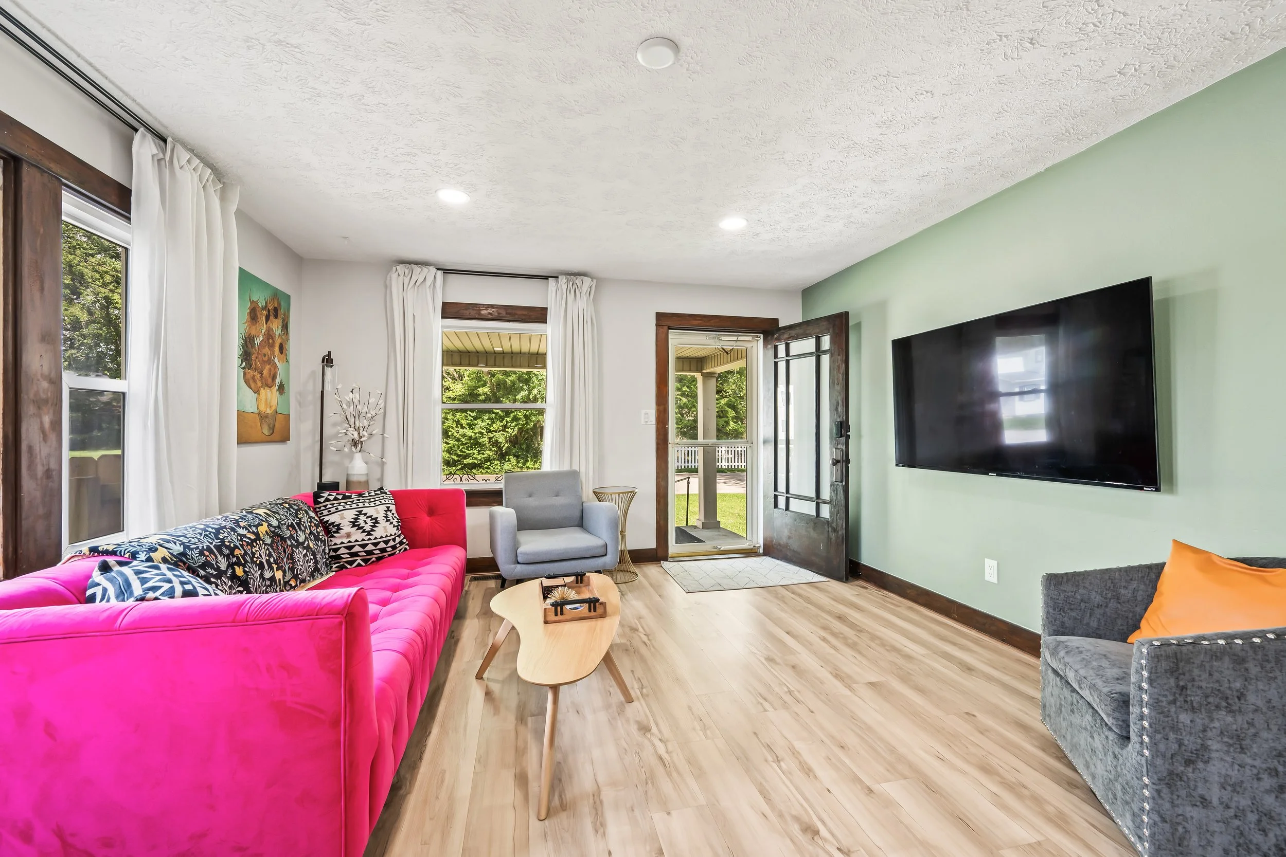A cozy living room with a pink velvet sofa, a gray armchair, and a small wooden coffee table. Large windows with white curtains reveal a green outdoor view. The room has hardwood floors, a light-colored wall with a painting, and a door leading outsid