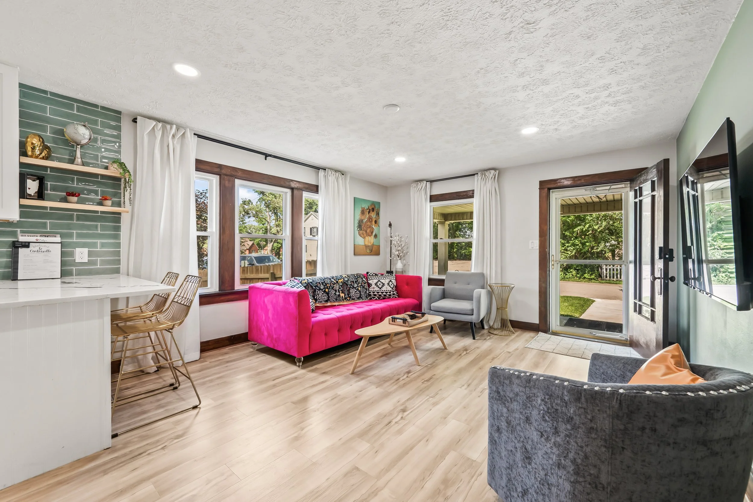 Living room with pink sofa, gray armchair, large windows with white curtains, and artwork of sunflowers on the wall.