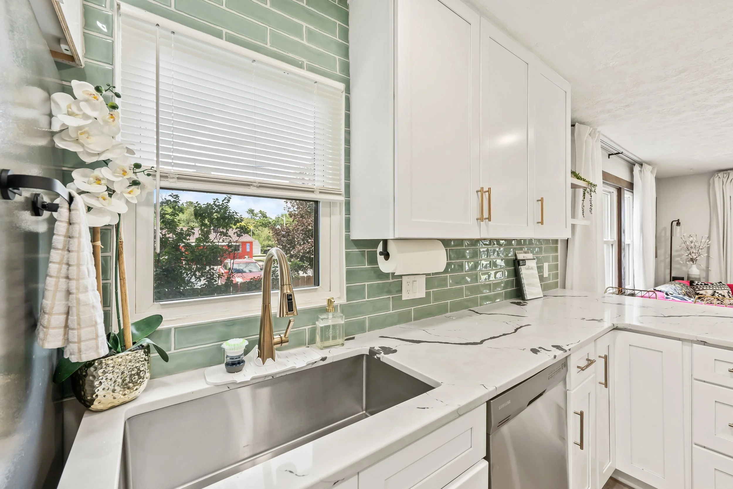 A modern kitchen with white cabinets, green subway tile backsplash, marble countertop, and a window with white blinds above the sink. Decor includes orchids, a soap dispenser, and kitchen towels.