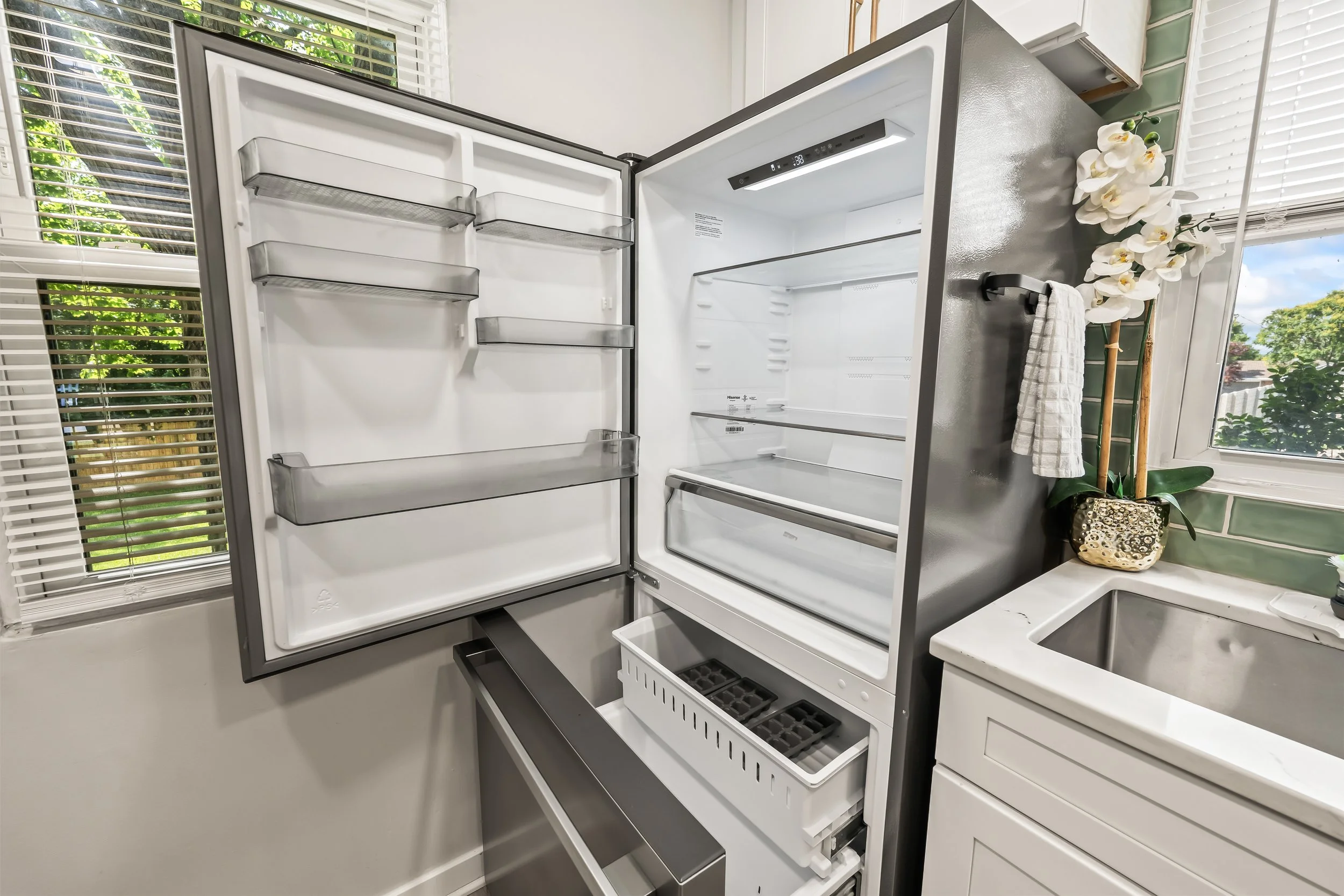 Empty modern refrigerator in a bright kitchen with white cabinets, a window with blinds, a vase of white orchids, and sunlit greenery outside.