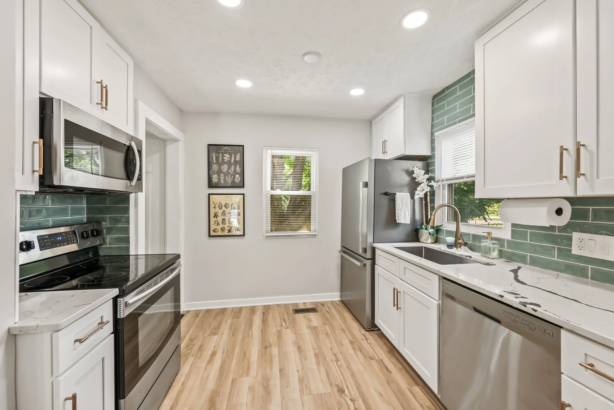 A modern kitchen with white cabinets, a marble countertop, green tile backsplash, stainless steel appliances including a microwave, oven, and refrigerator, and two windows with white blinds.