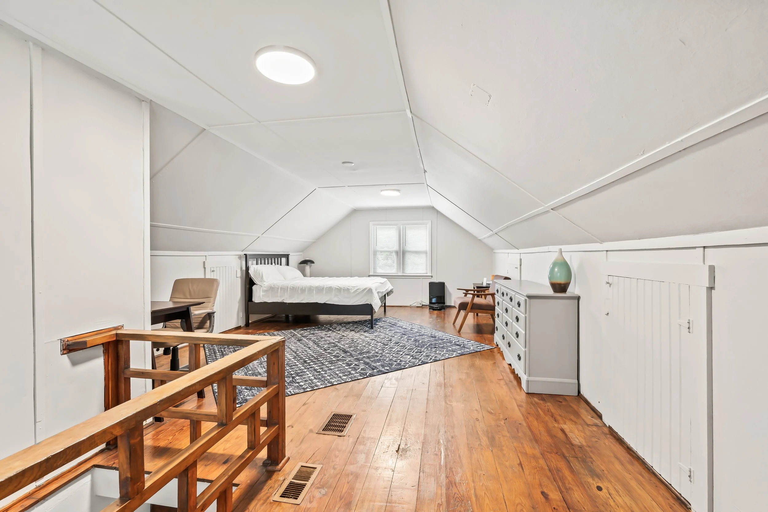 A spacious loft bedroom with white walls and ceiling, hardwood floors, a bed near the window, a large dresser with a decorative vase, a seating area with chairs, and a patterned rug.