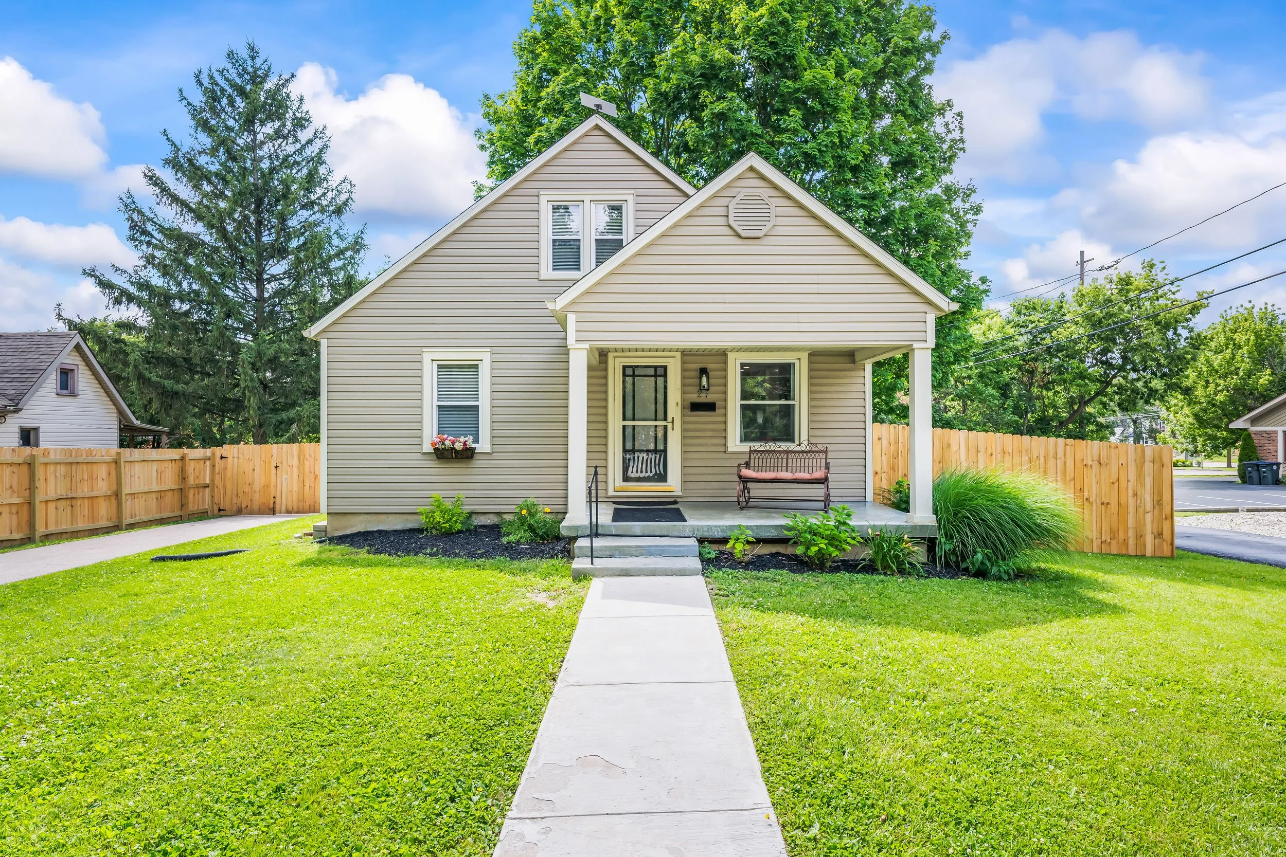 A beige two-story house with a small front porch, surrounded by a green lawn and a wooden fence. The house has two windows on the front, a door with a screen, and a bench on the porch. There are trees in the background under a partly cloudy sky.