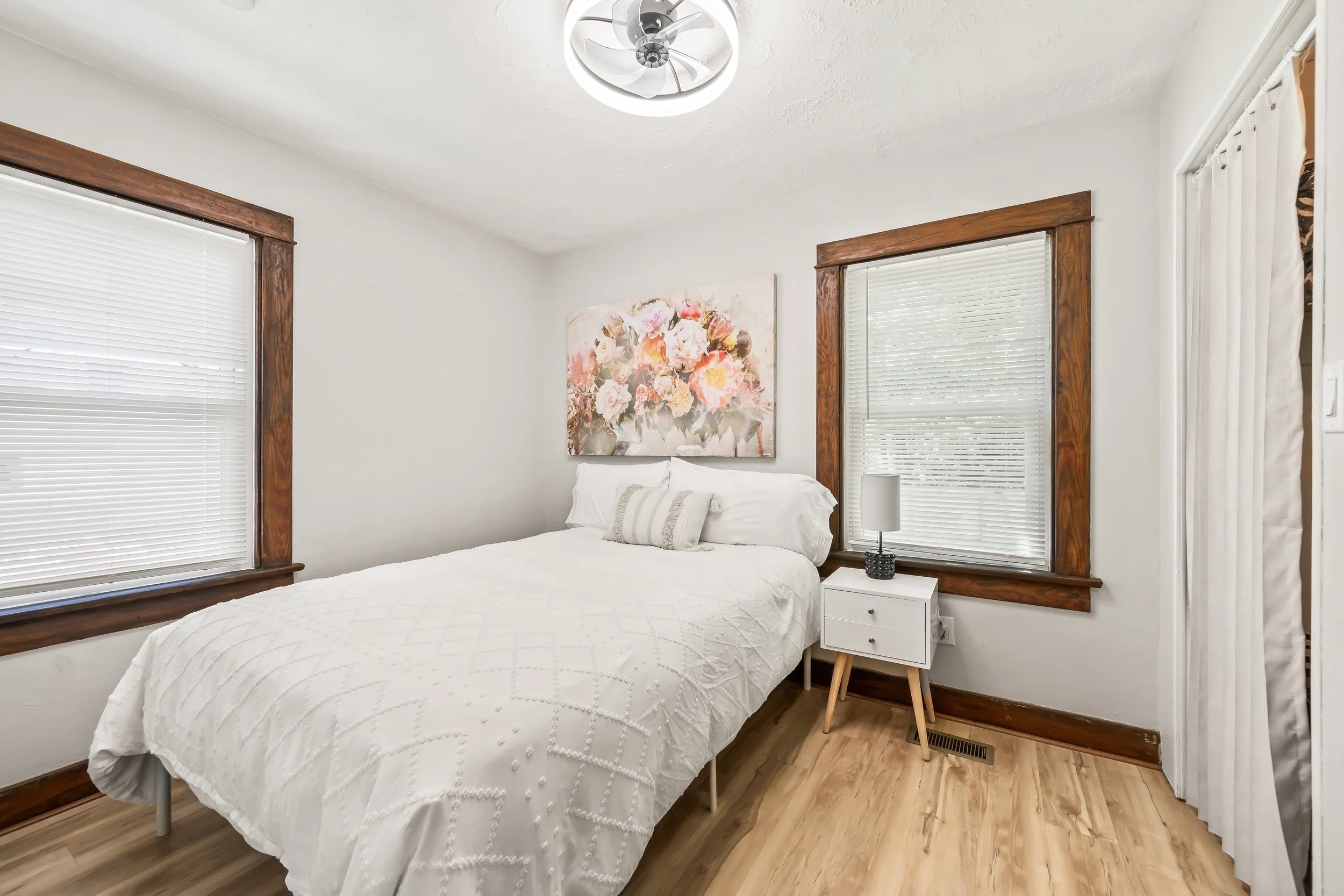 A bedroom with a bed, white bedding, and pillows, two windows with white blinds, framed with dark wood, a chandelier on the ceiling, a framed floral painting above the bed, a bedside table with a lamp, and a wooden chair.