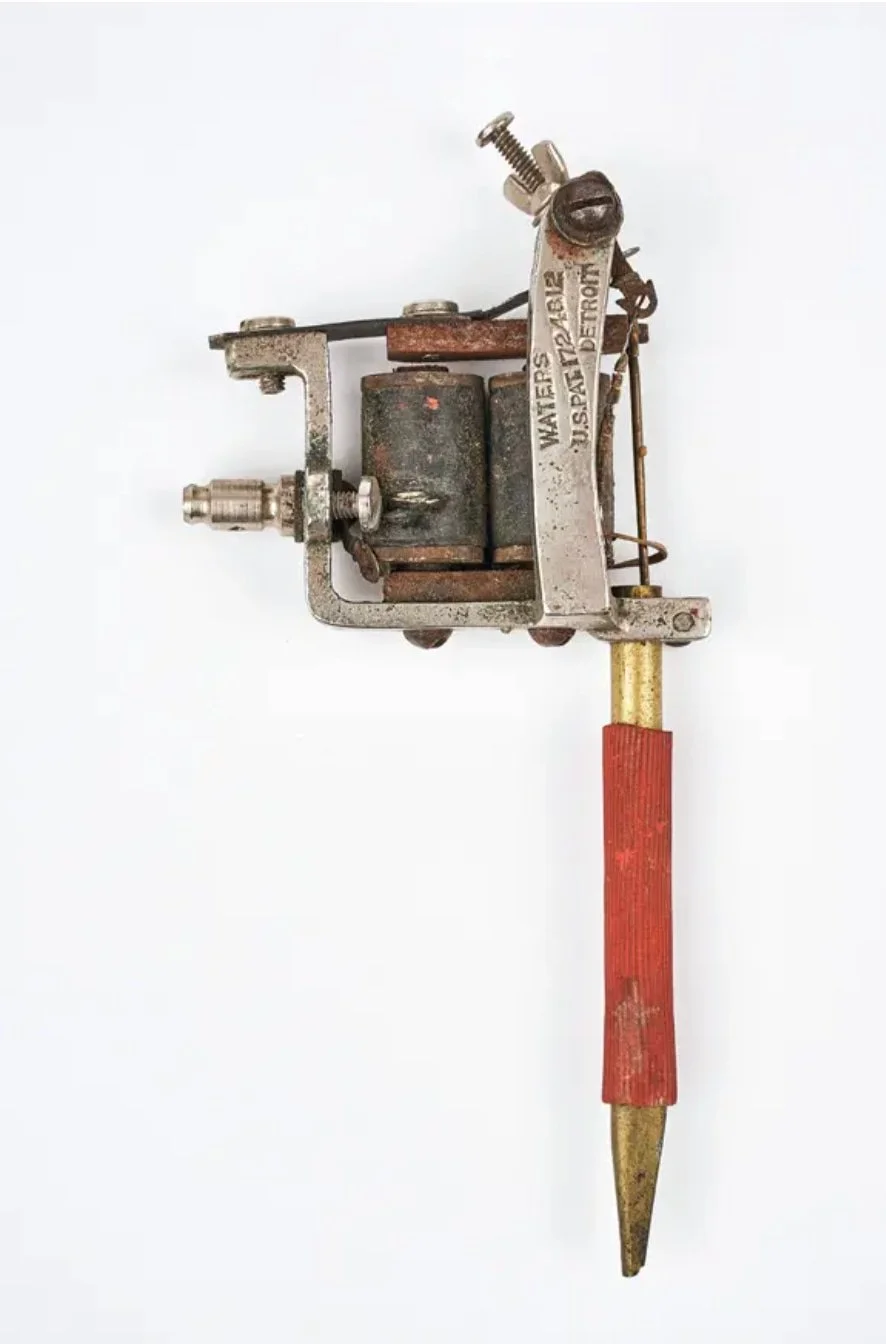 A vintage tattoo machine with a red grip, metal frame, and visible coils against a white background.