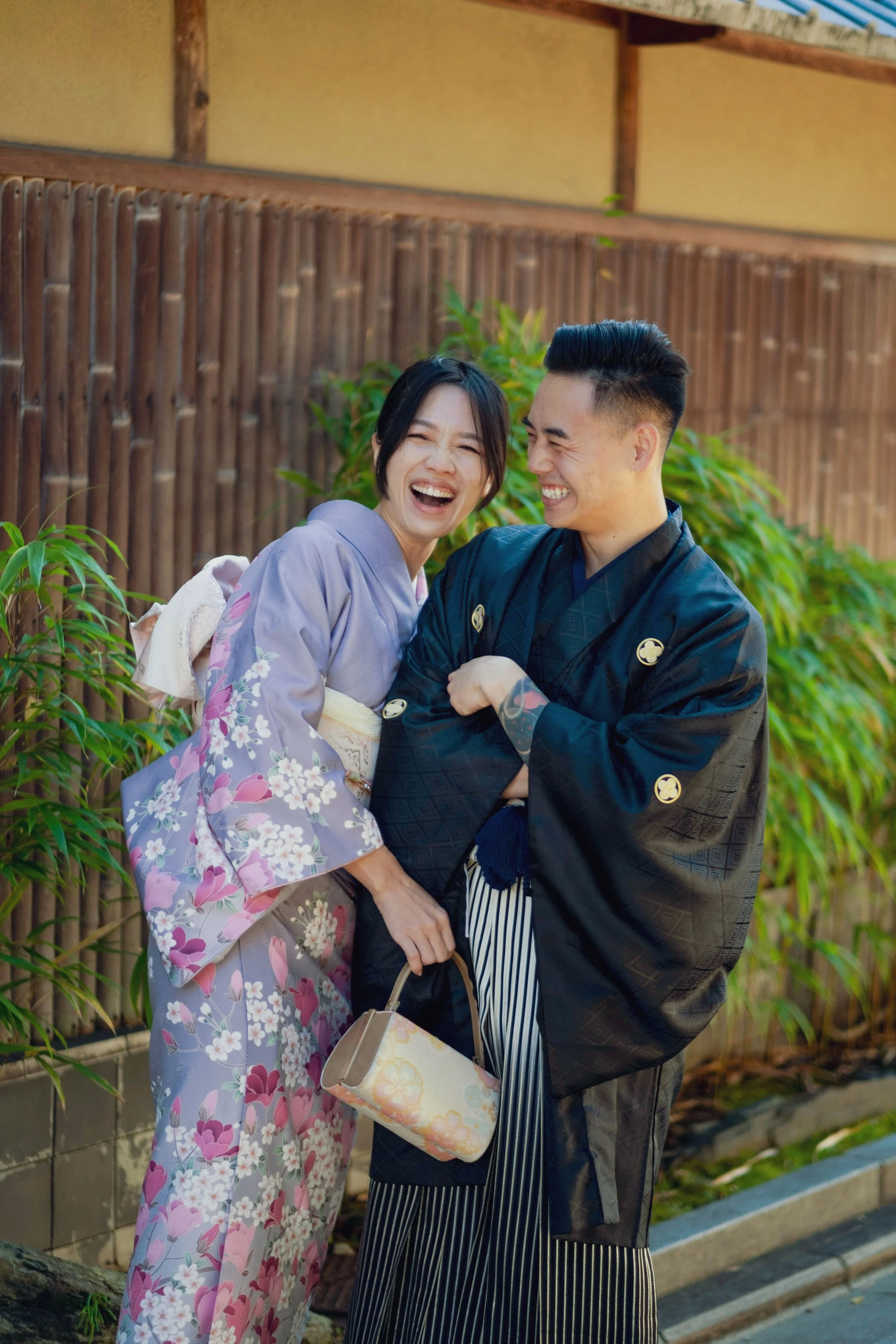 A smiling woman and man in traditional Japanese kimonos sharing a joyful moment outdoors, standing in front of a bamboo fence and green foliage.