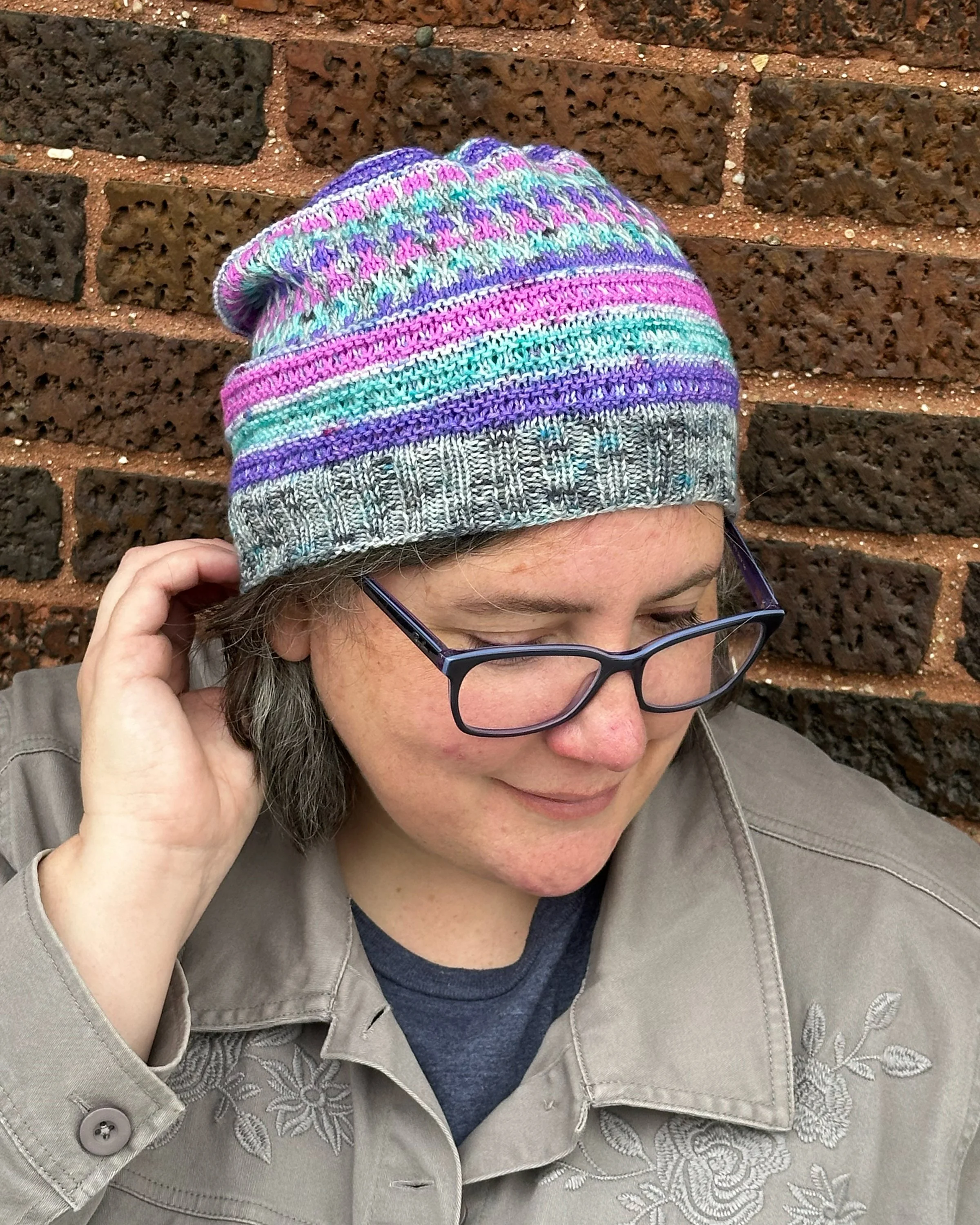 Middle-aged white woman modeling a brightly colored striped knitted hat, standing in front of a brick wall.