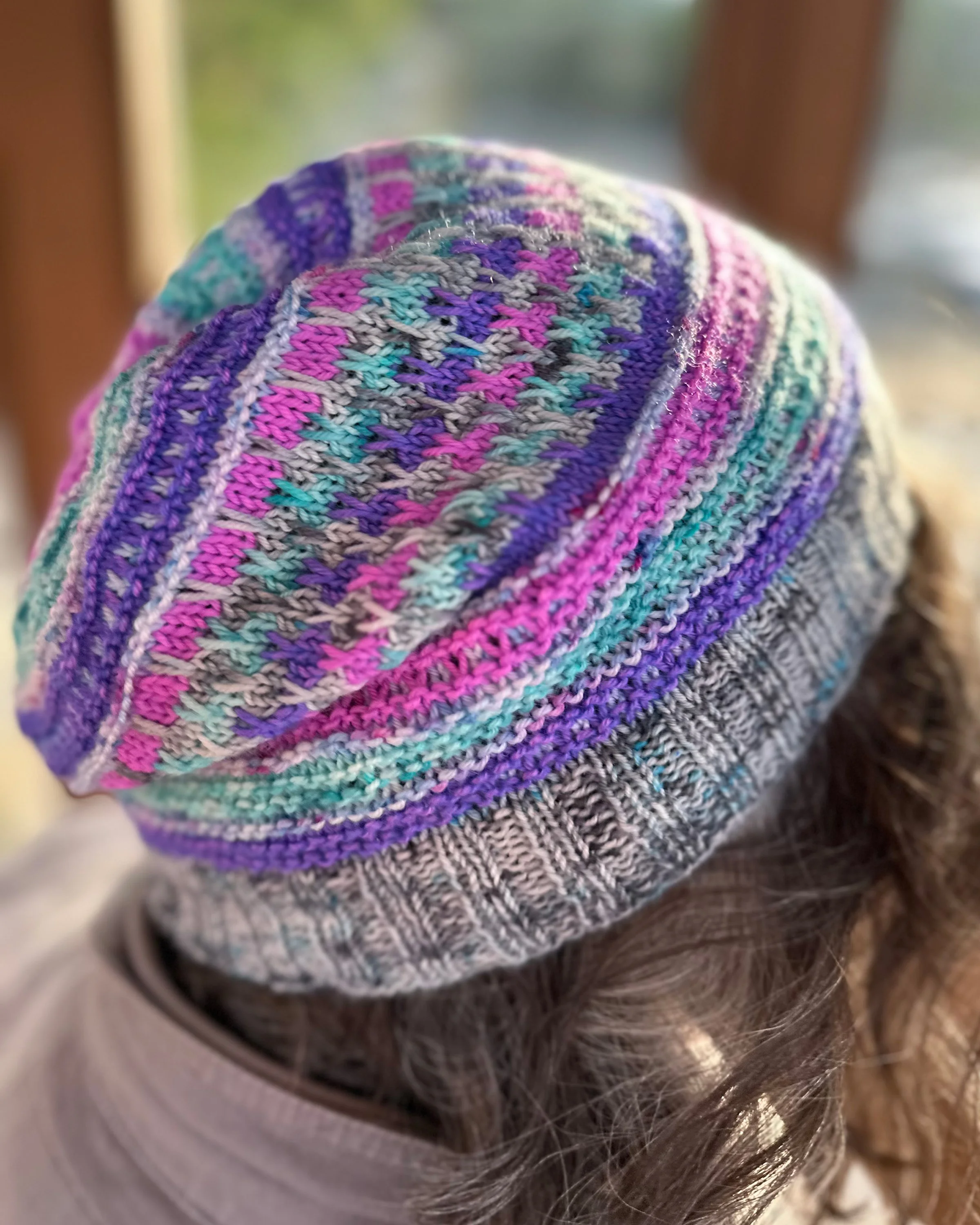 Close up photo of a the side of a woman's head, modeling a slipped and garter stitch knitted hat, worked in bright colors.