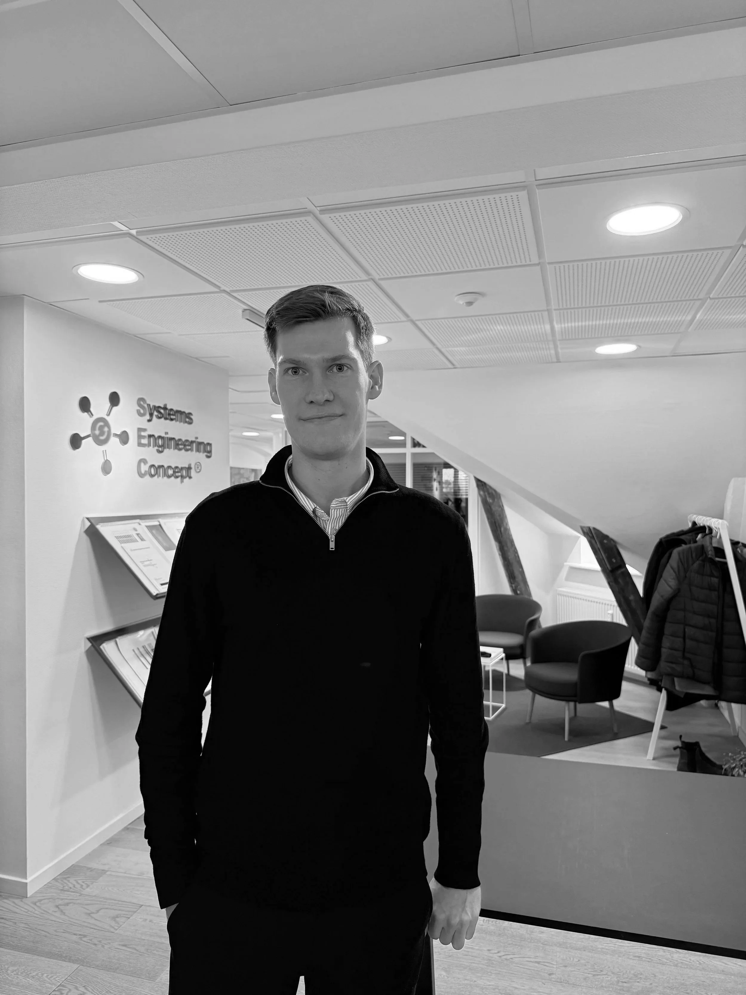 A young man standing indoors in front of a wall with a logo and text reading 'Systems Engineering Concept.' The setting appears to be an office or conference space with chairs and a coat rack in the background.