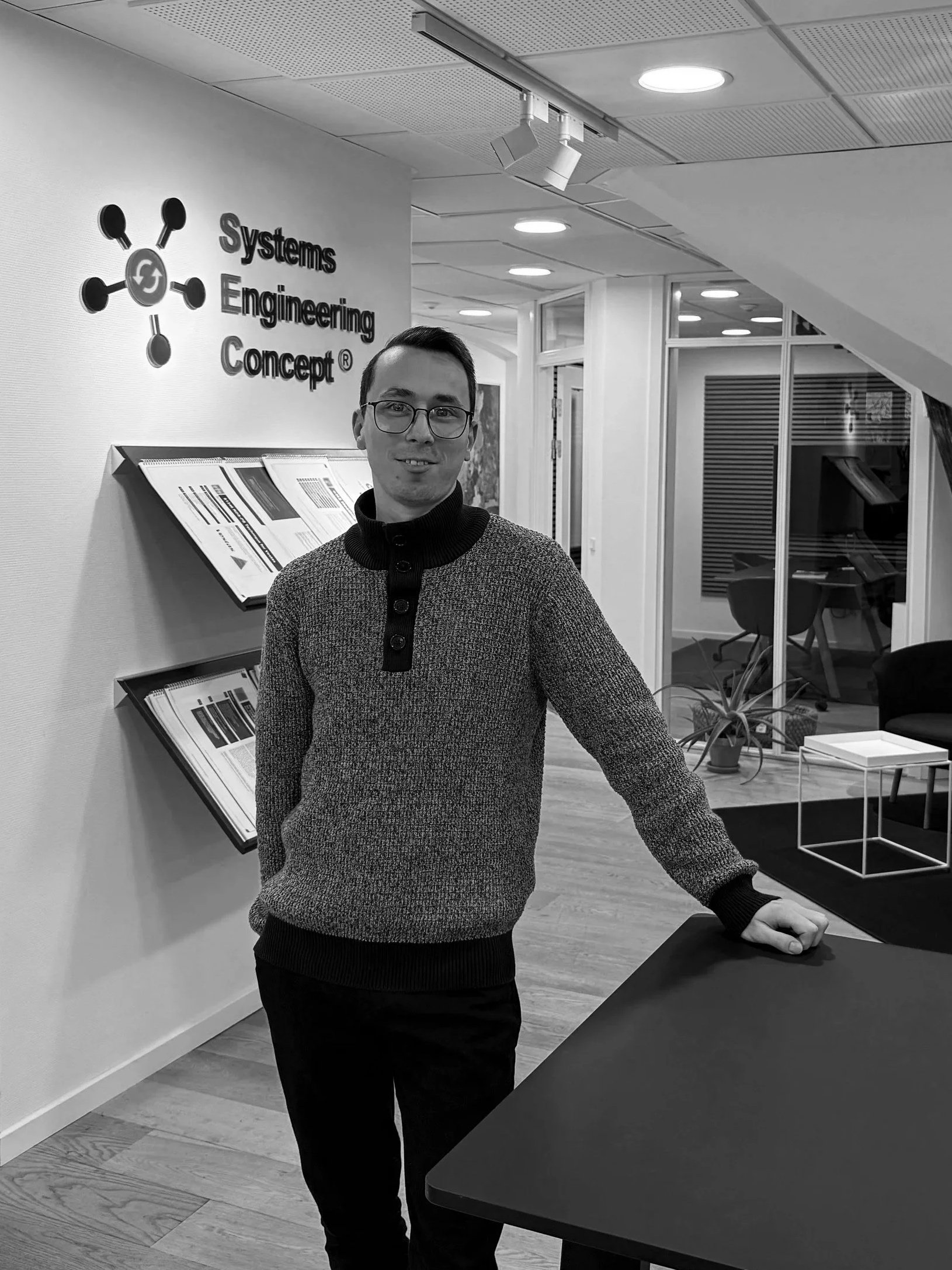 A man standing in an office space, leaning on a table, with the logo and name 'Systems Engineering Concept' on the wall behind him.