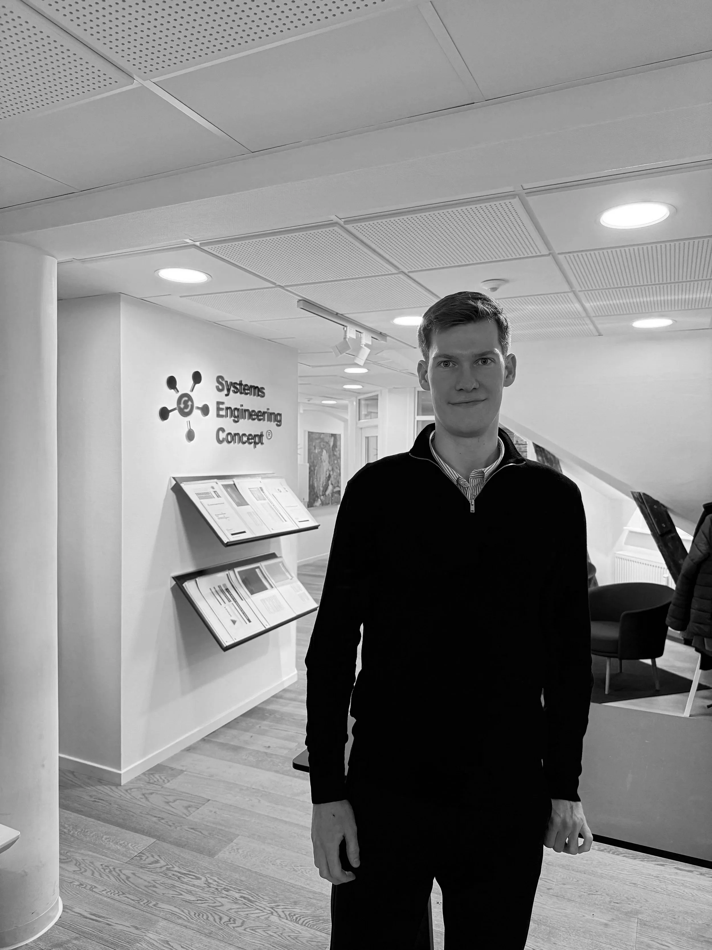A young man standing indoors in front of a wall with a logo and text reading 'Systems Engineering Concept.' The setting appears to be an office or conference space with chairs and a coat rack in the background.