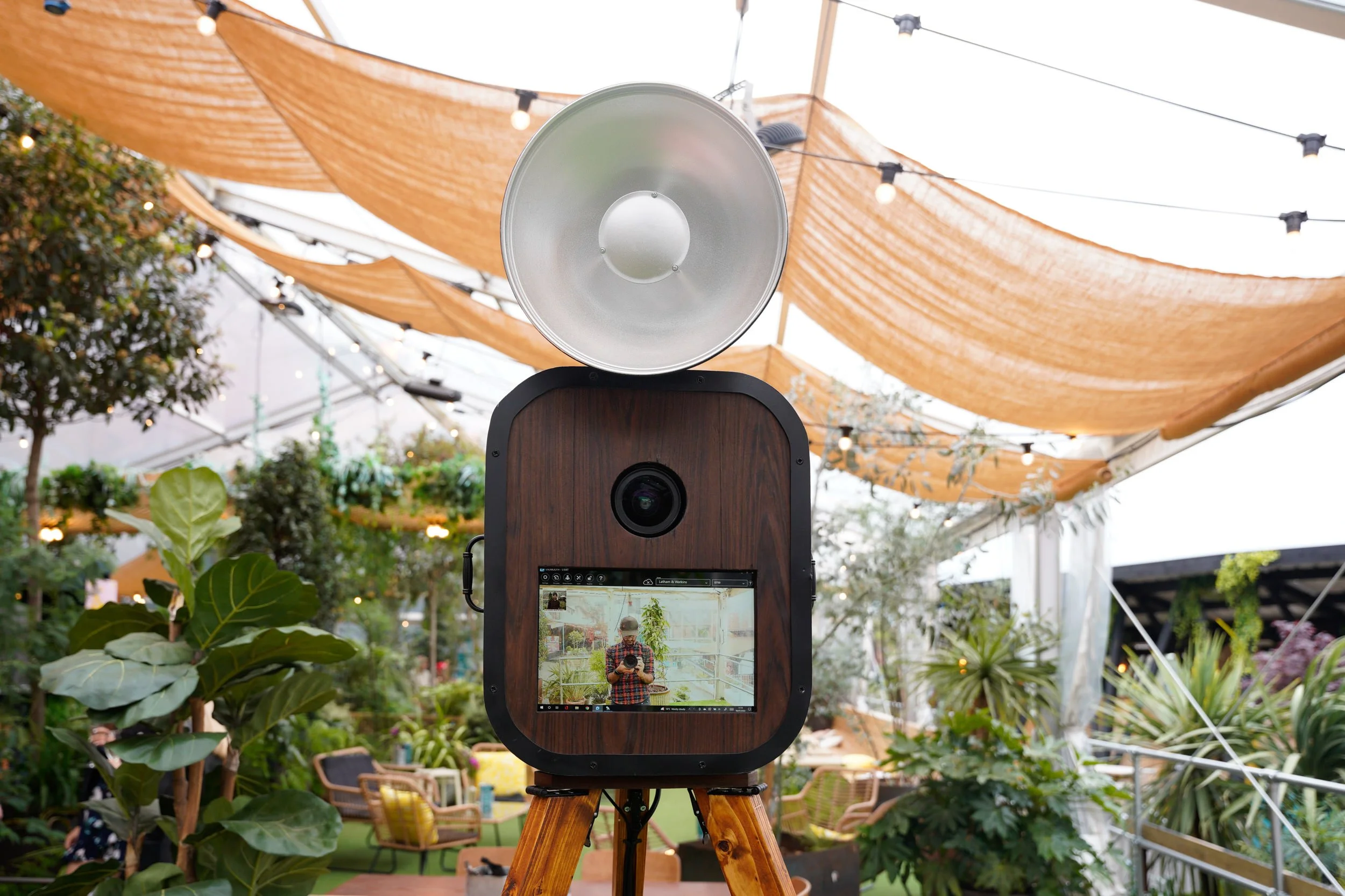 A vintage-style camera on a tripod capturing a person taking a photo of the camera inside a lush indoor garden with orange fabric canopies and string lights.