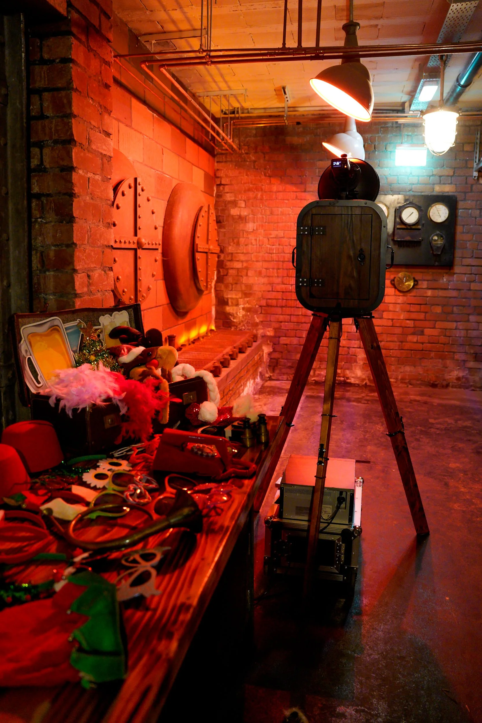 Vintage camera on tripod pointing toward a brick wall with retro industrial wall decor, warm red lighting, and miscellaneous props and decorations on a wooden table.