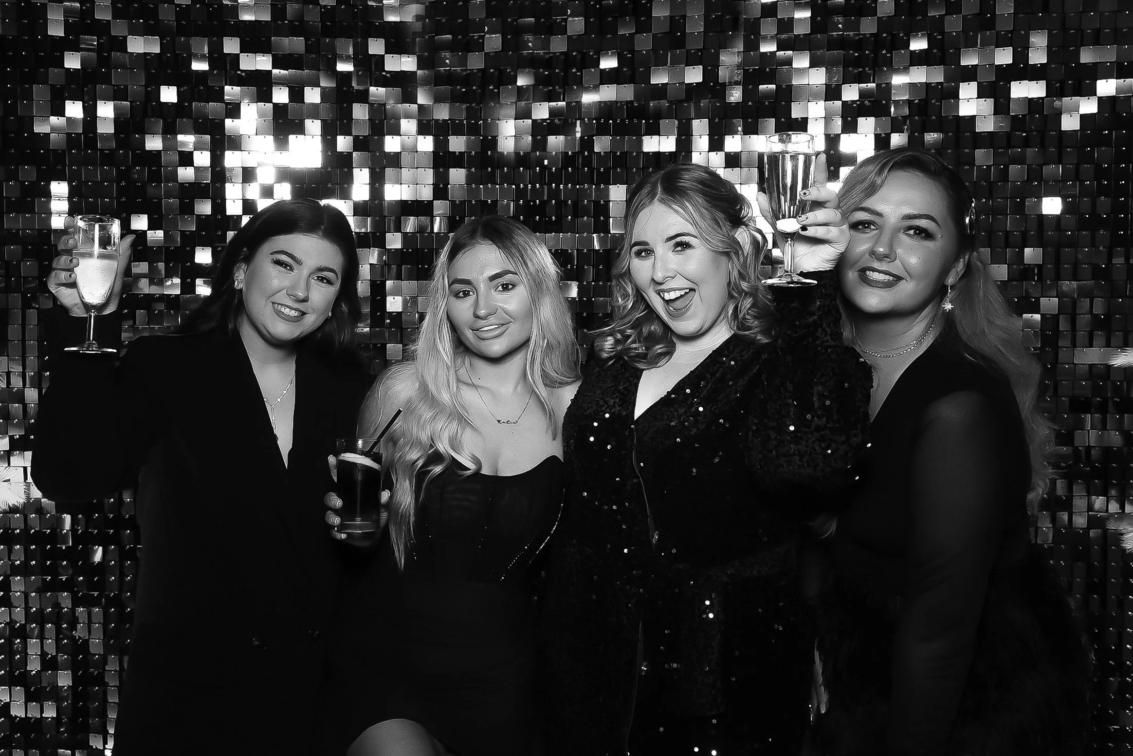 Four women celebrating at a party, smiling and holding glasses of champagne, with a sparkly, sequined backdrop.