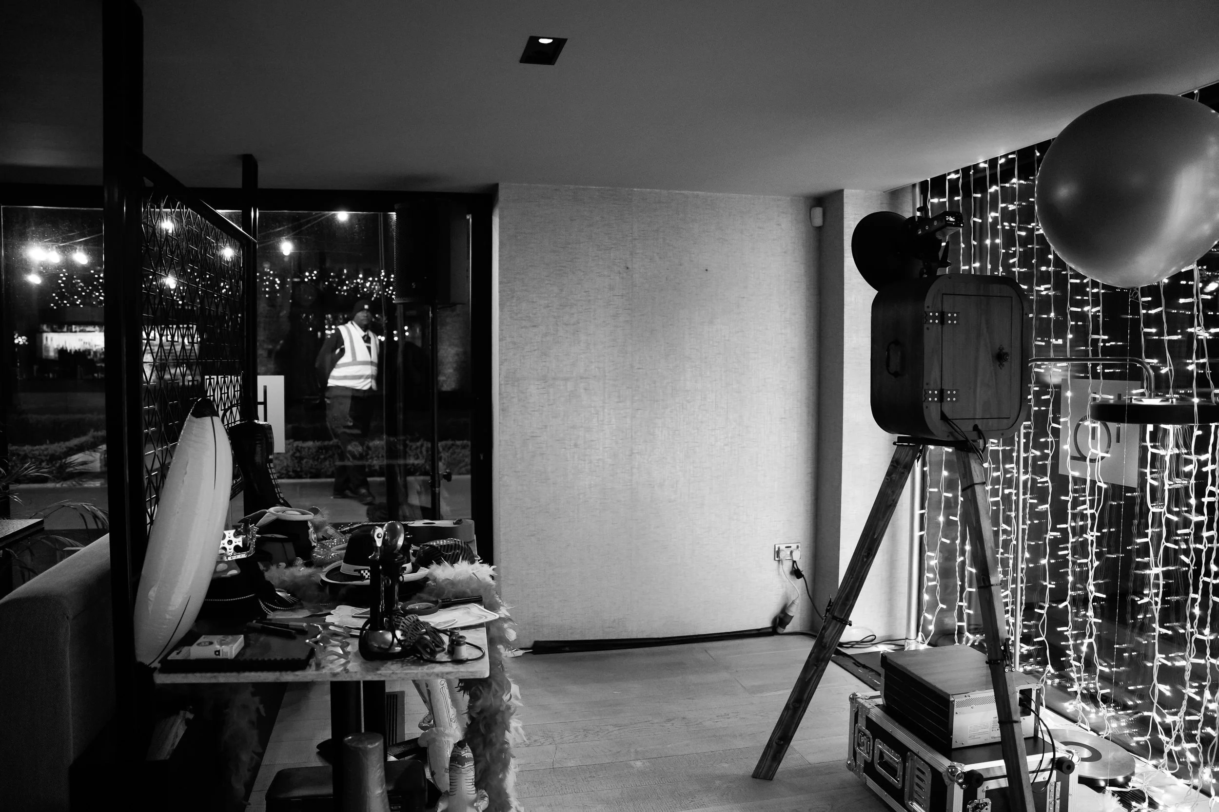 A black and white indoor scene with a camera on a tripod, string lights on the walls, a large balloon, and a table cluttered with various items, near a glass door through which a person is visible outside.