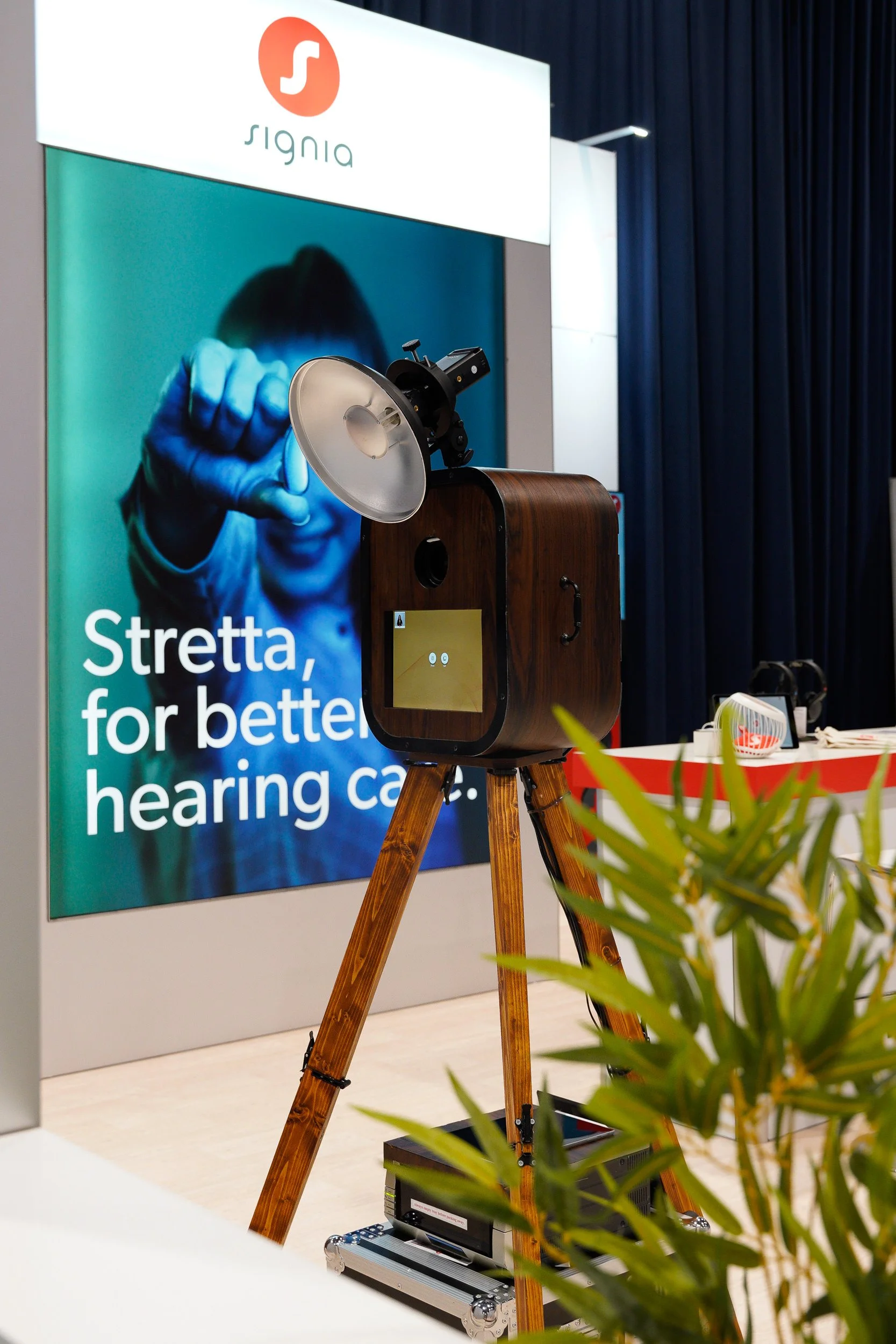 A modern hearing test device or audiometer on a wooden tripod stand at an exhibition. In the background, there is a large banner with the logo and the text 'Stretta, for better hearing care.' A plant partially obscures the view in the foreground.
