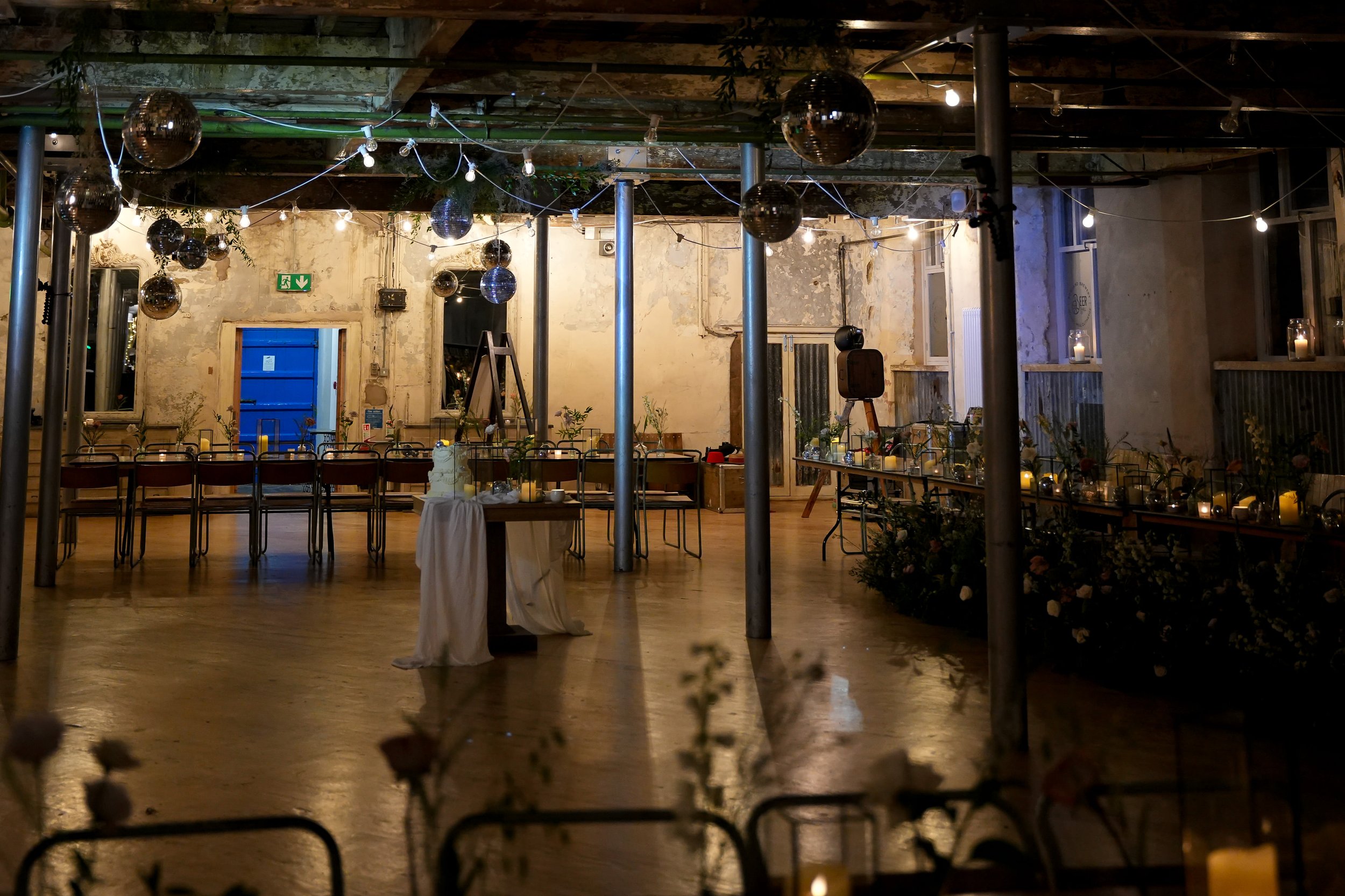 A decorated event space with string lights and disco balls hanging from the ceiling, tables arranged for a celebration, floral arrangements, candles, and a rustic, slightly worn wall appearance.