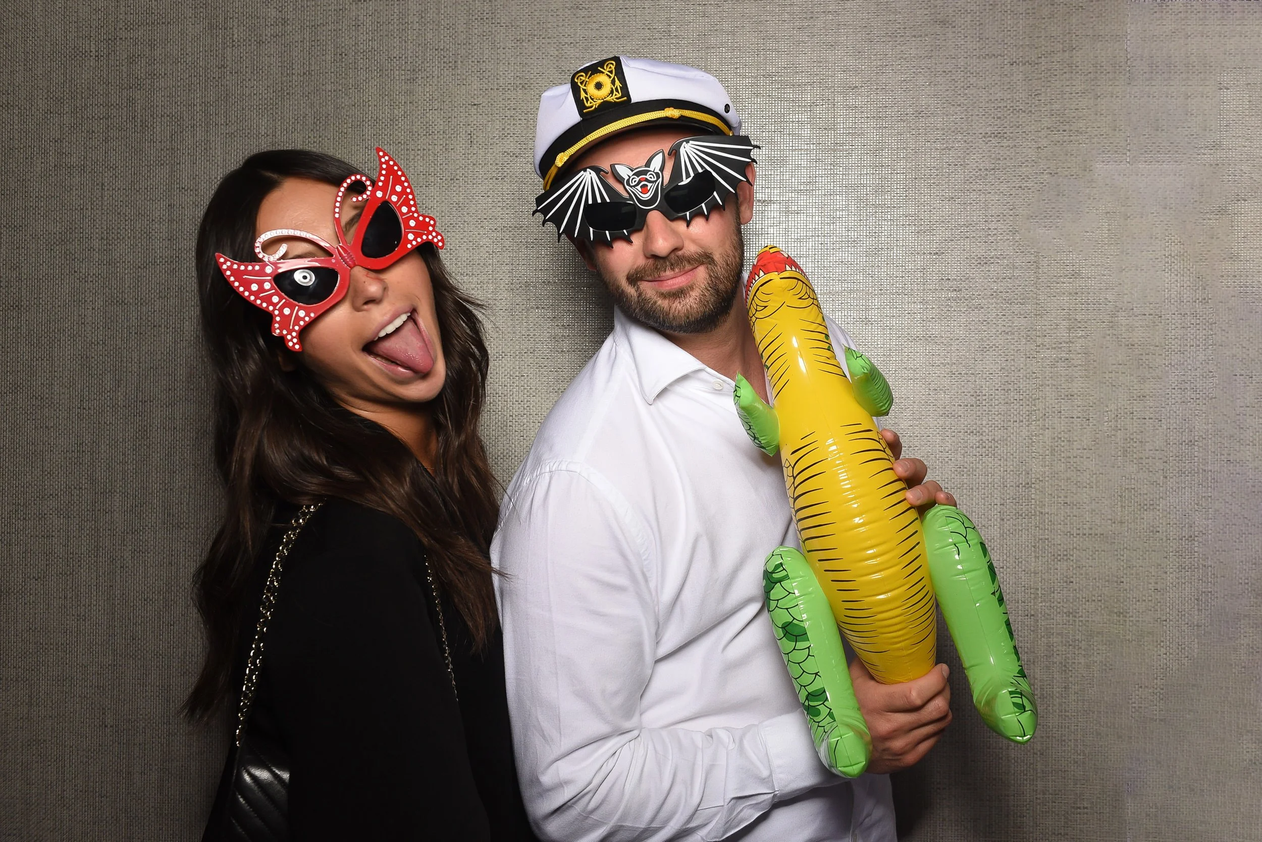 Two people wearing fun masks and accessories, one woman with a red butterfly mask sticking out her tongue, and one man with a gorilla mask, a captain's hat, and holding an inflatable crocodile.