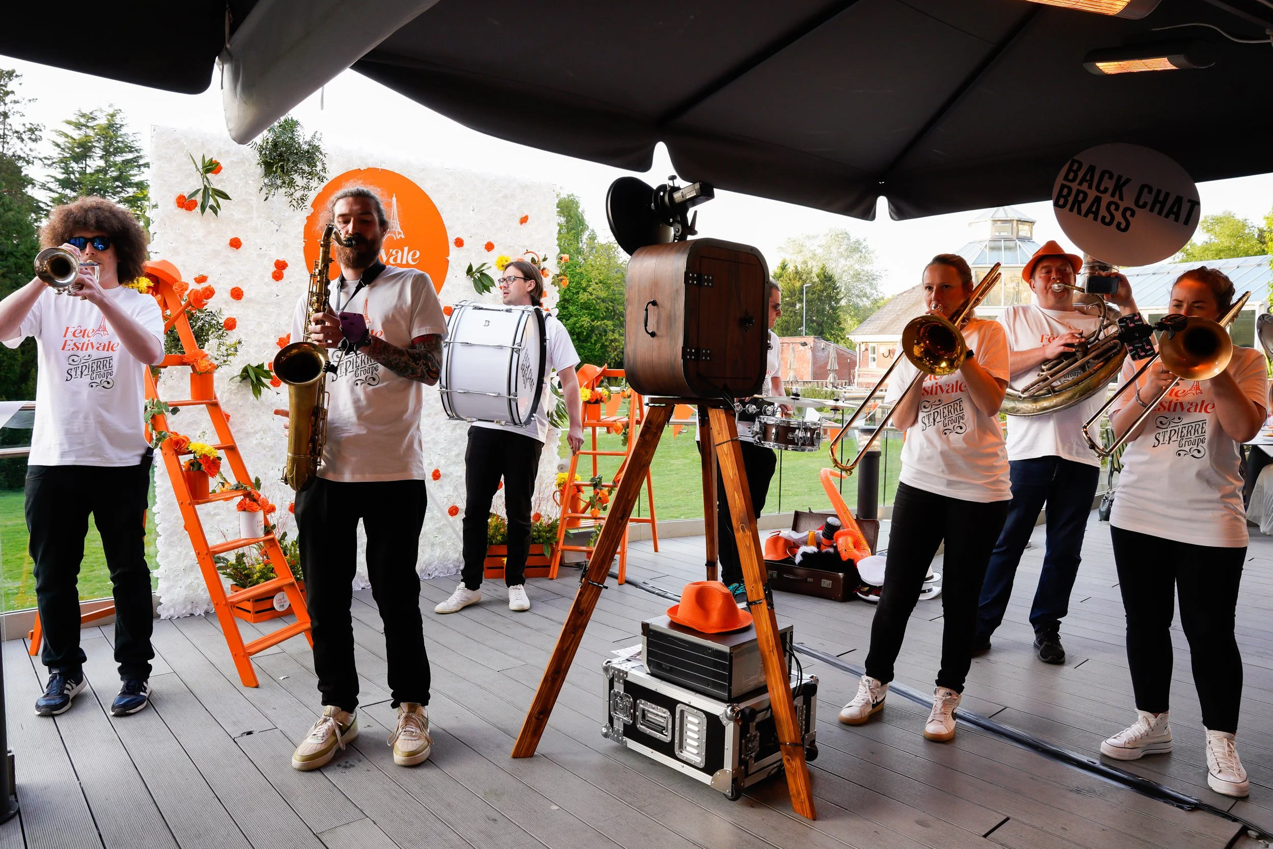 A band of six people playing instruments on an outdoor stage at a festival, with a white backdrop featuring an orange logo and orange decorations.
