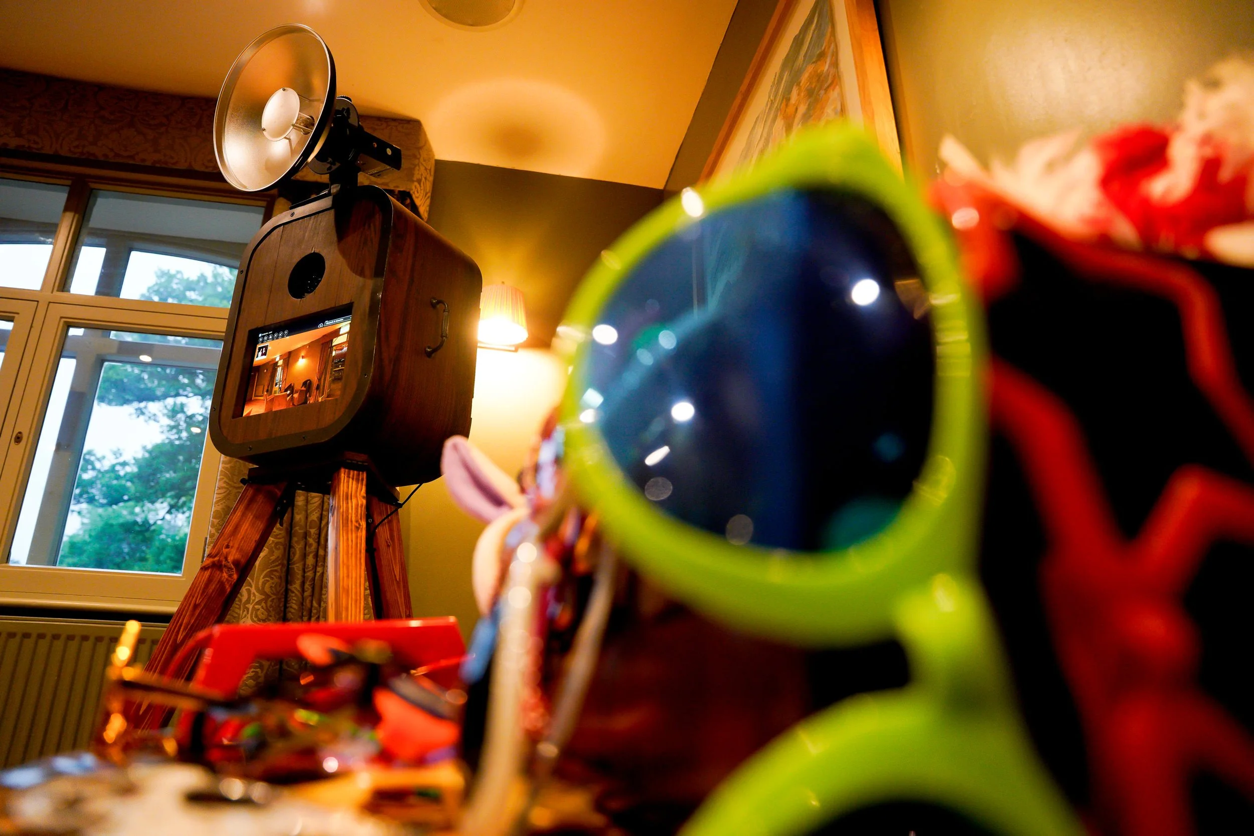 Close-up of colorful sunglasses on a table in front of a vintage wooden camera on a tripod inside a room with windows and framed artwork.