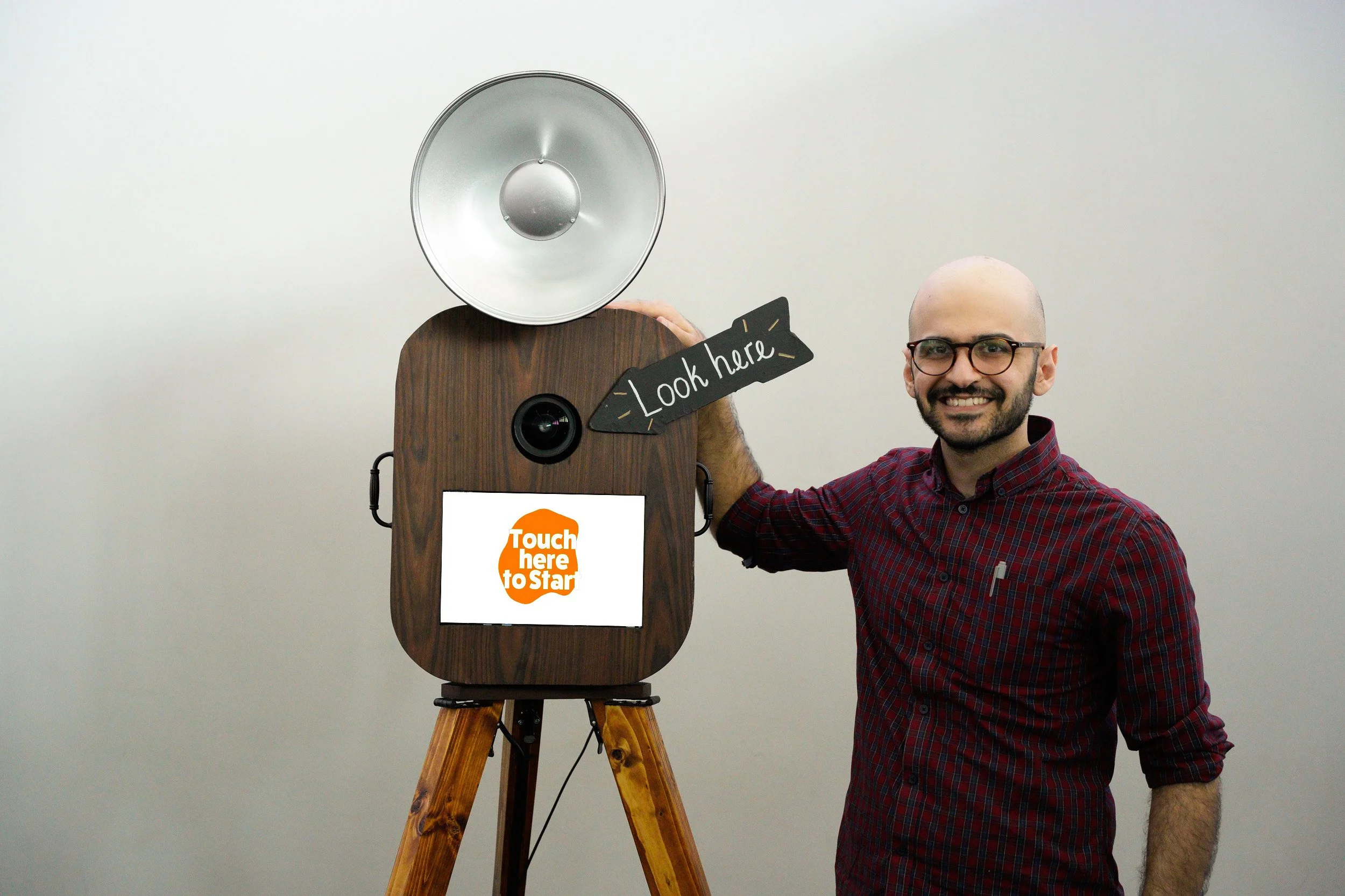 A man with glasses and a red checkered shirt standing next to an interactive photo booth with a sign that says 'Look here' and a screen that says 'Touch here to Star.' The booth has a camera, a large silver spotlight, and is mounted on wooden tripod 