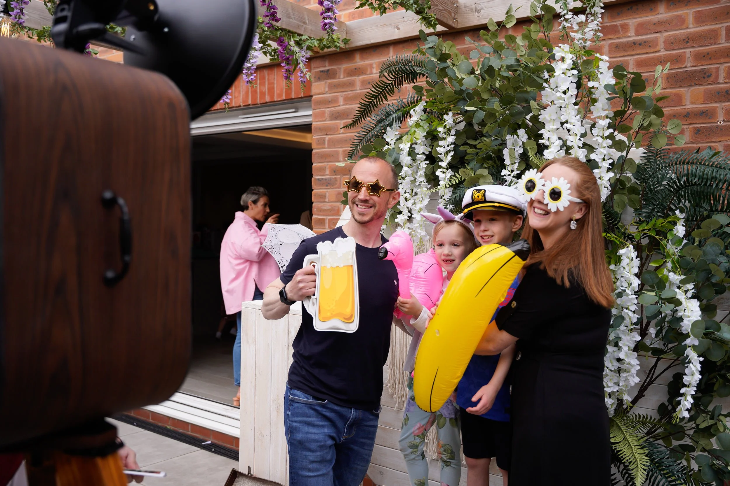 Group of four people dressed in fun costumes celebrating outdoors in front of a floral backdrop, with a camera in the foreground capturing the moment.