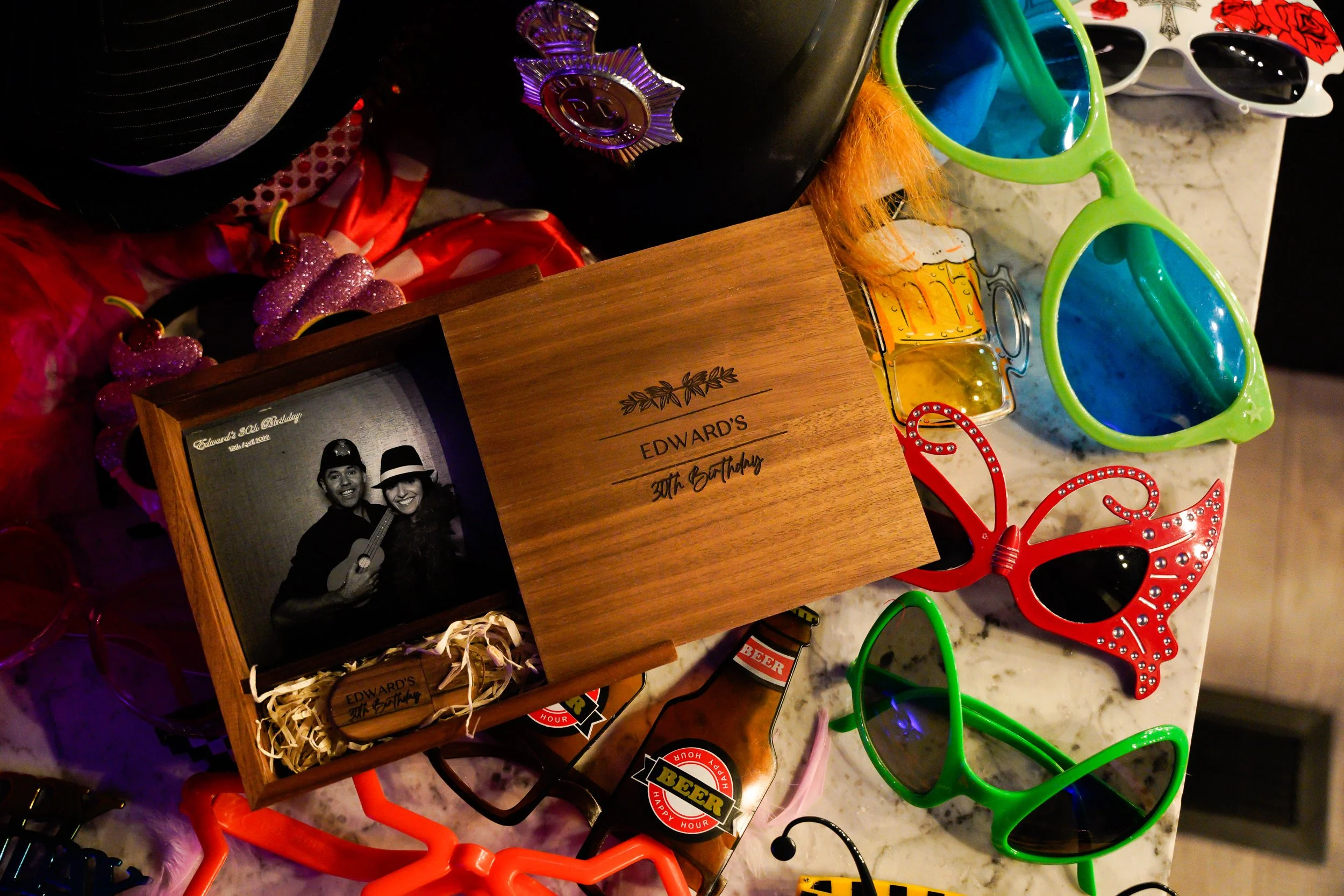 A birthday celebration display including a wooden box with a black and white photo of two people, colorful sunglasses, bottles of beer shaped mini liquor bottles, and decorative items. The box has an inscription that reads 'Edward's 30th Birthday.'