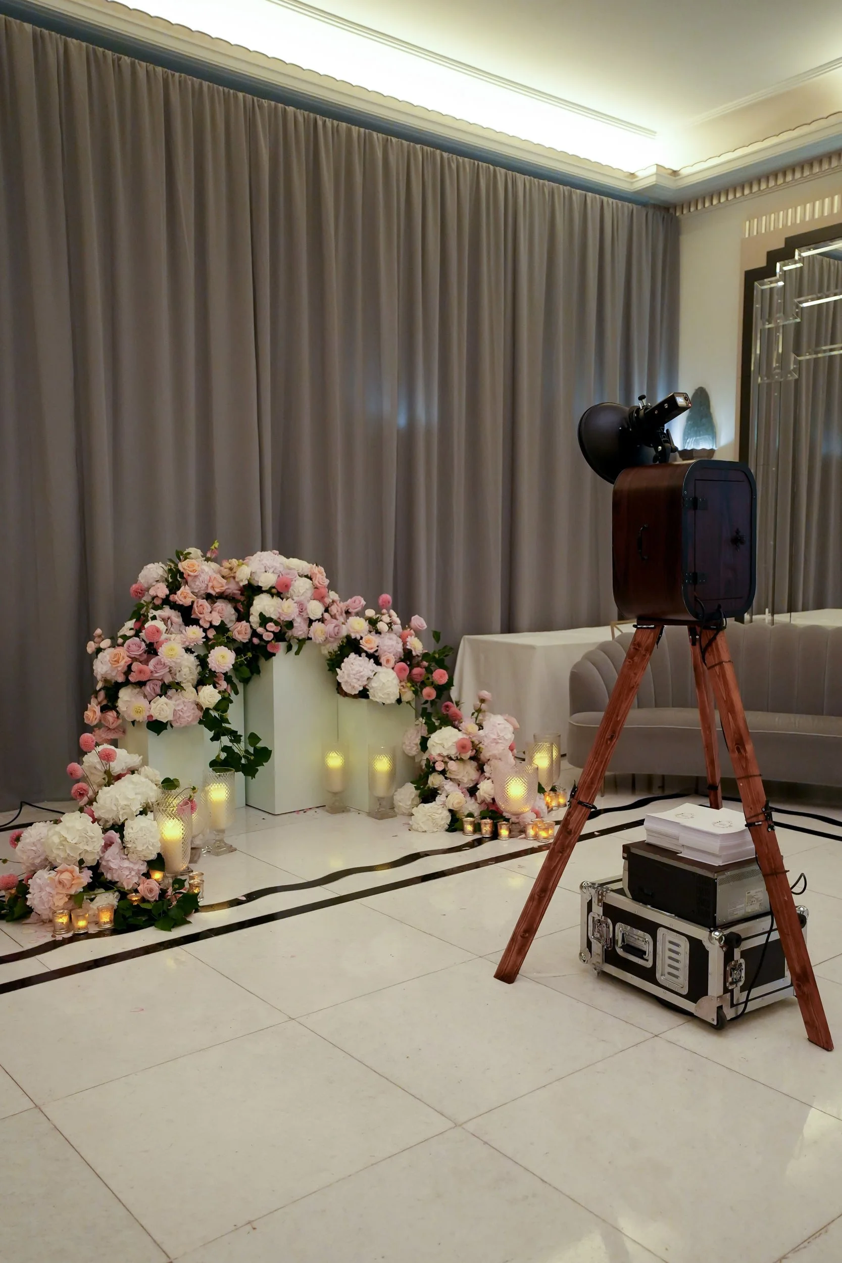 Wedding or event decoration setup with a floral arrangement of pink and white roses, hydrangeas, candles, and lanterns arranged on the floor in front of a beige curtain. A photo booth or camera with equipment is set up nearby.