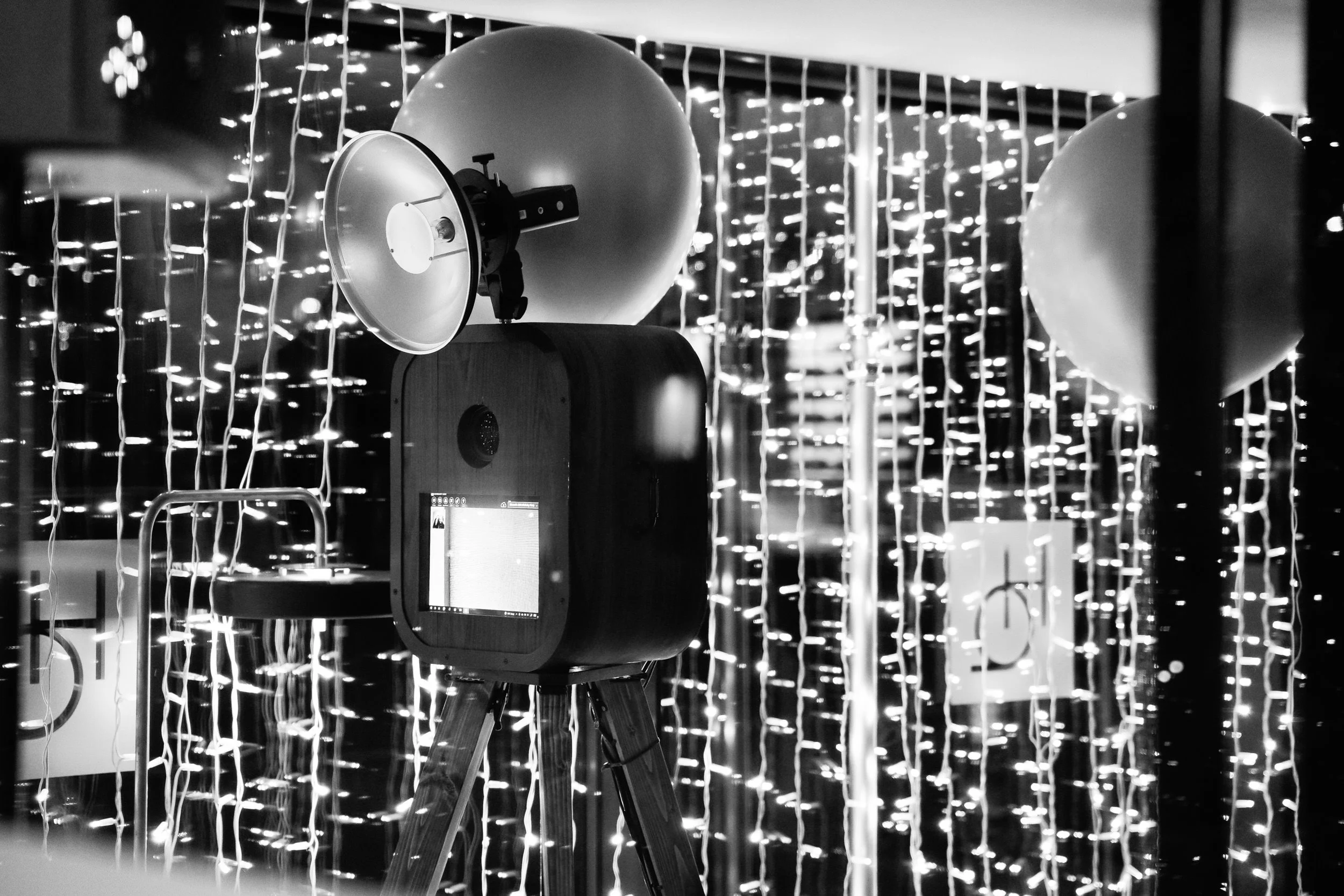 A vintage film camera on a tripod in front of hanging string lights and balloons, black and white photo.