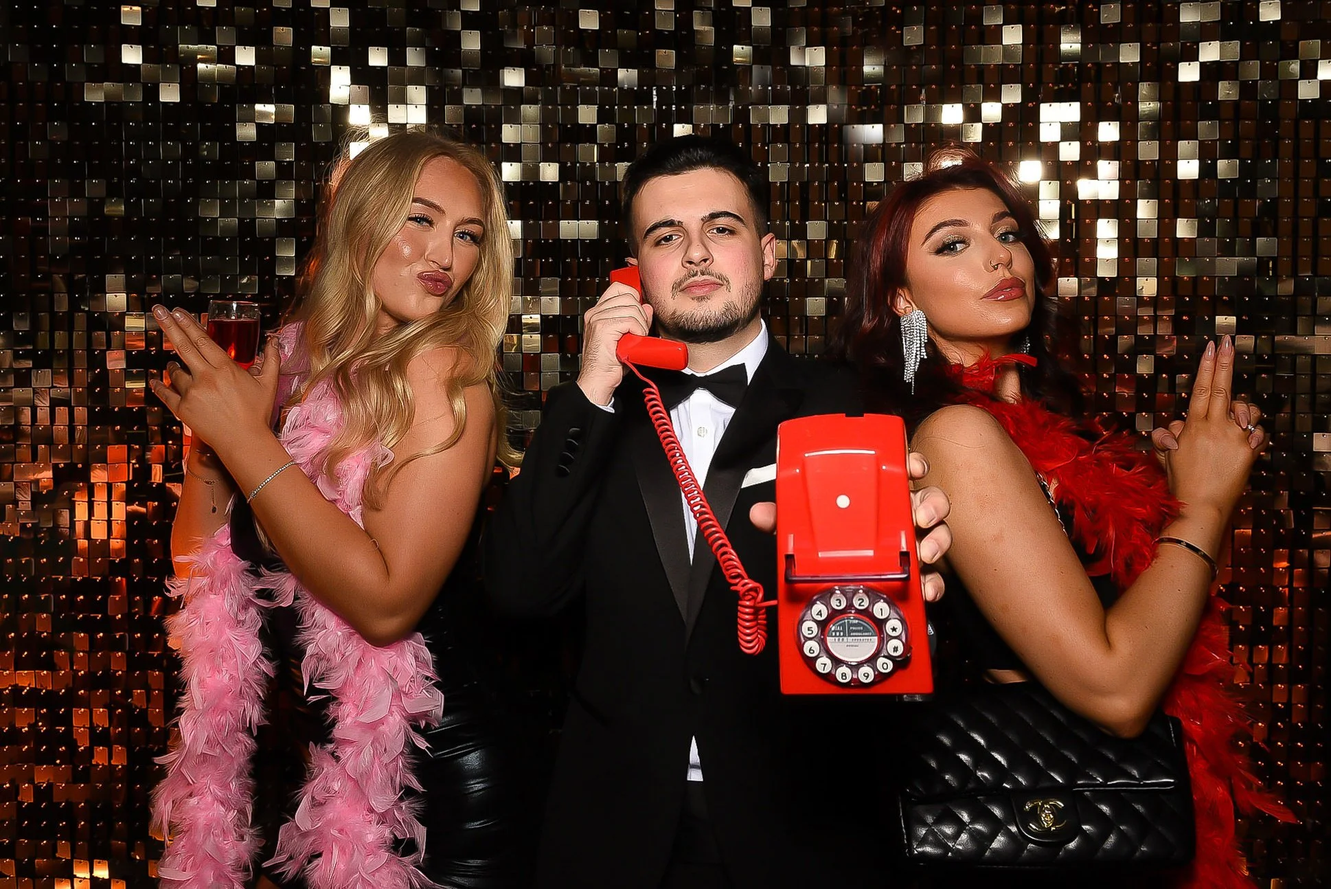Three people dressed in formal attire posing against a shiny, sequin backdrop, with two women wearing feather boas and a man in a tuxedo holding a vintage red rotary phone.