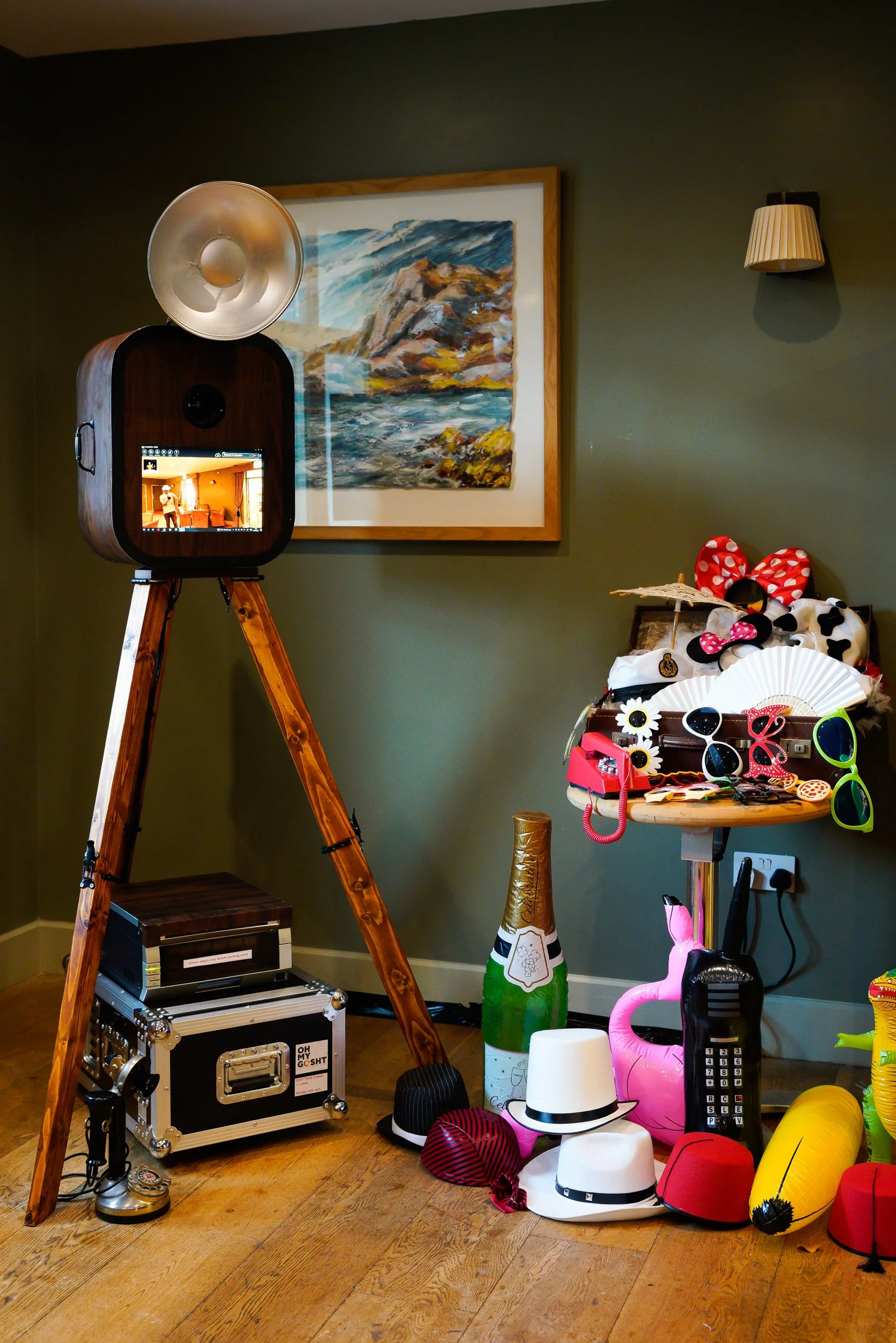 A collection of party props and decorations including hats, sunglasses, a flamingo float, a champagne bottle or prop, and a telephone, arranged next to a wall with a painting, in a room with wooden flooring.