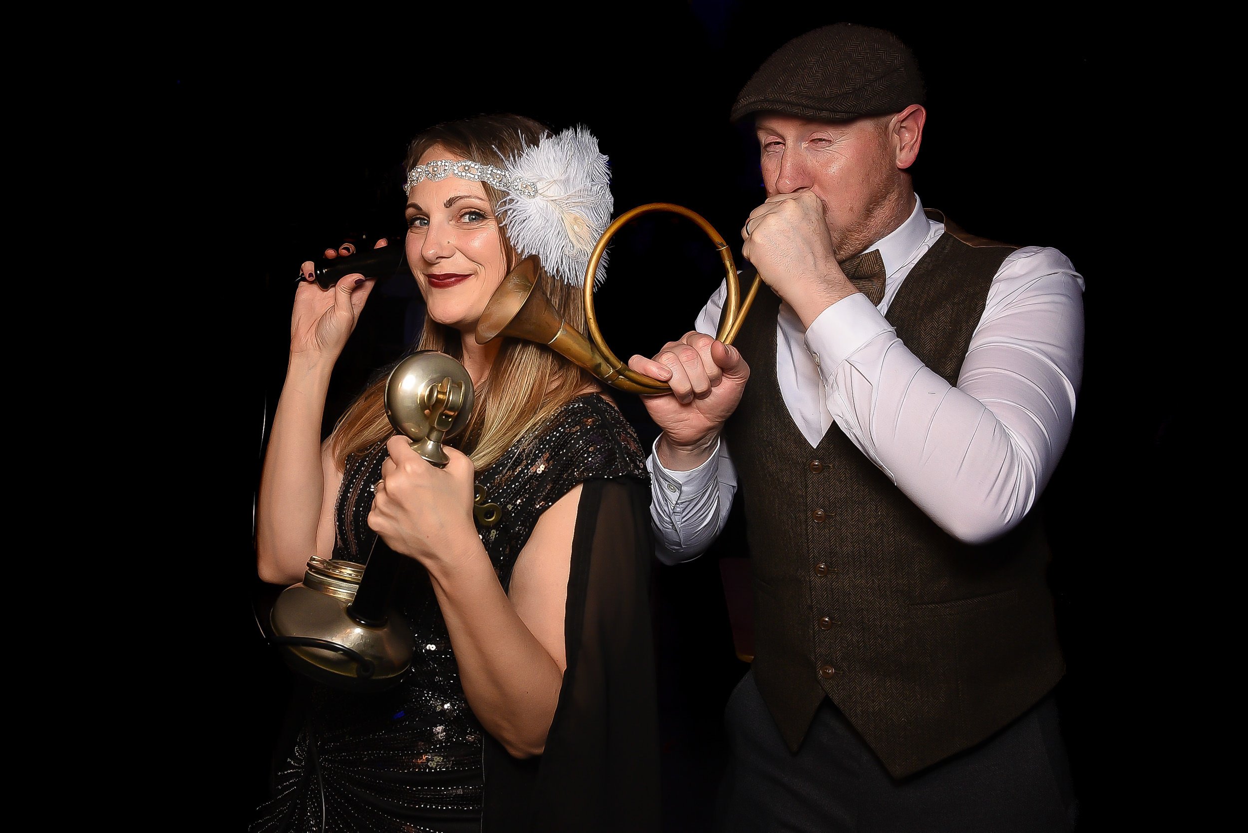 A man and woman dressed in vintage 1920s style clothing playing musical instruments. The woman is holding an old-fashioned telephone receiver and a small trumpet, while the man is playing a horn. The woman has a feather headband and the man is wearin